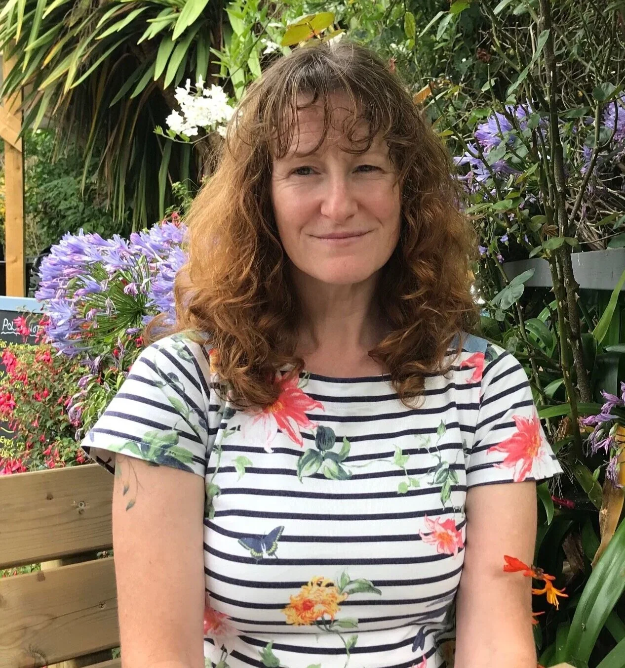 A woman with curly, shoulder-length hair sitting outdoors among flowering plants, wearing a striped floral t-shirt.