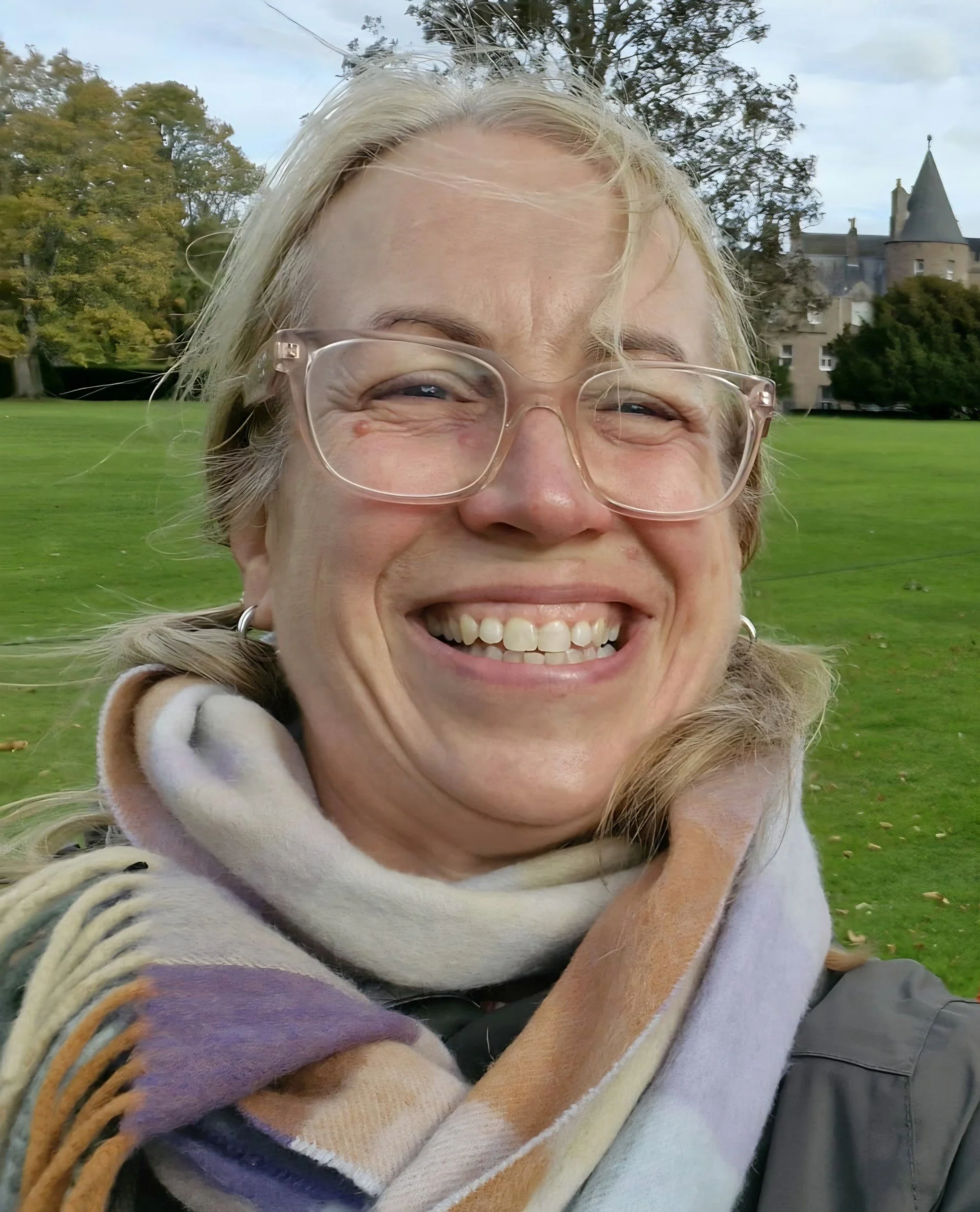Close-up of a smiling woman with glasses, wearing a multicolored scarf, outdoors on a grassy field with trees and a castle-like building in the background.