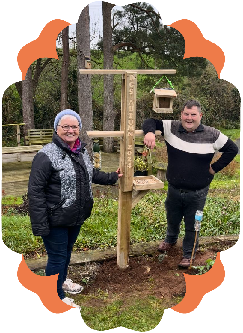 A woman and a man standing next to a wooden bird feeder with the words "TGS Autumn 2024" engraved on it, outdoors in a wooded area.
