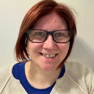 A woman with short red hair wearing black glasses, smiling, against a plain light-colored wall.