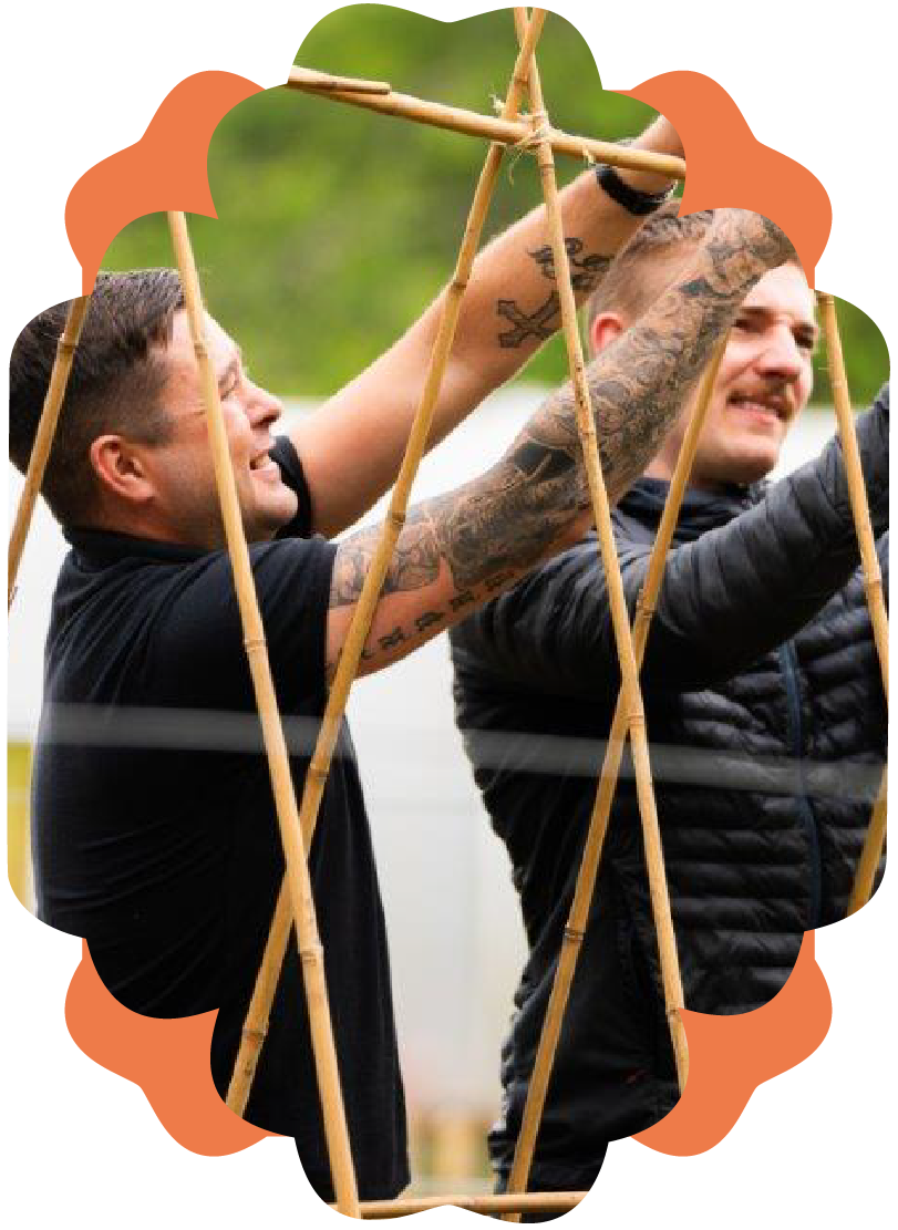 Two men with tattoos building a bamboo structure outdoors, smiling and working together.