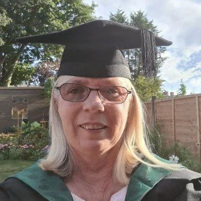 Woman wearing graduation gown and cap, smiling outdoors in a backyard with a wooden fence and trees.