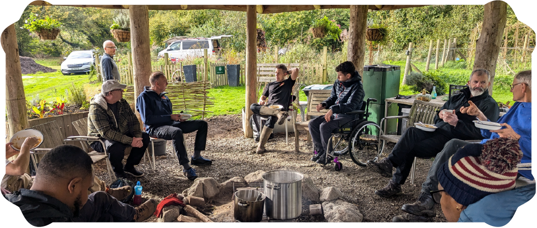 A group of diverse people sitting and chatting outdoors under a wooden shelter with a fire pit in the center, surrounded by trees and greenery.