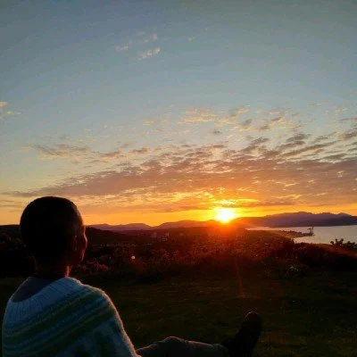 Person sitting outdoors, watching a sunset over a landscape with a body of water and mountains in the distance.