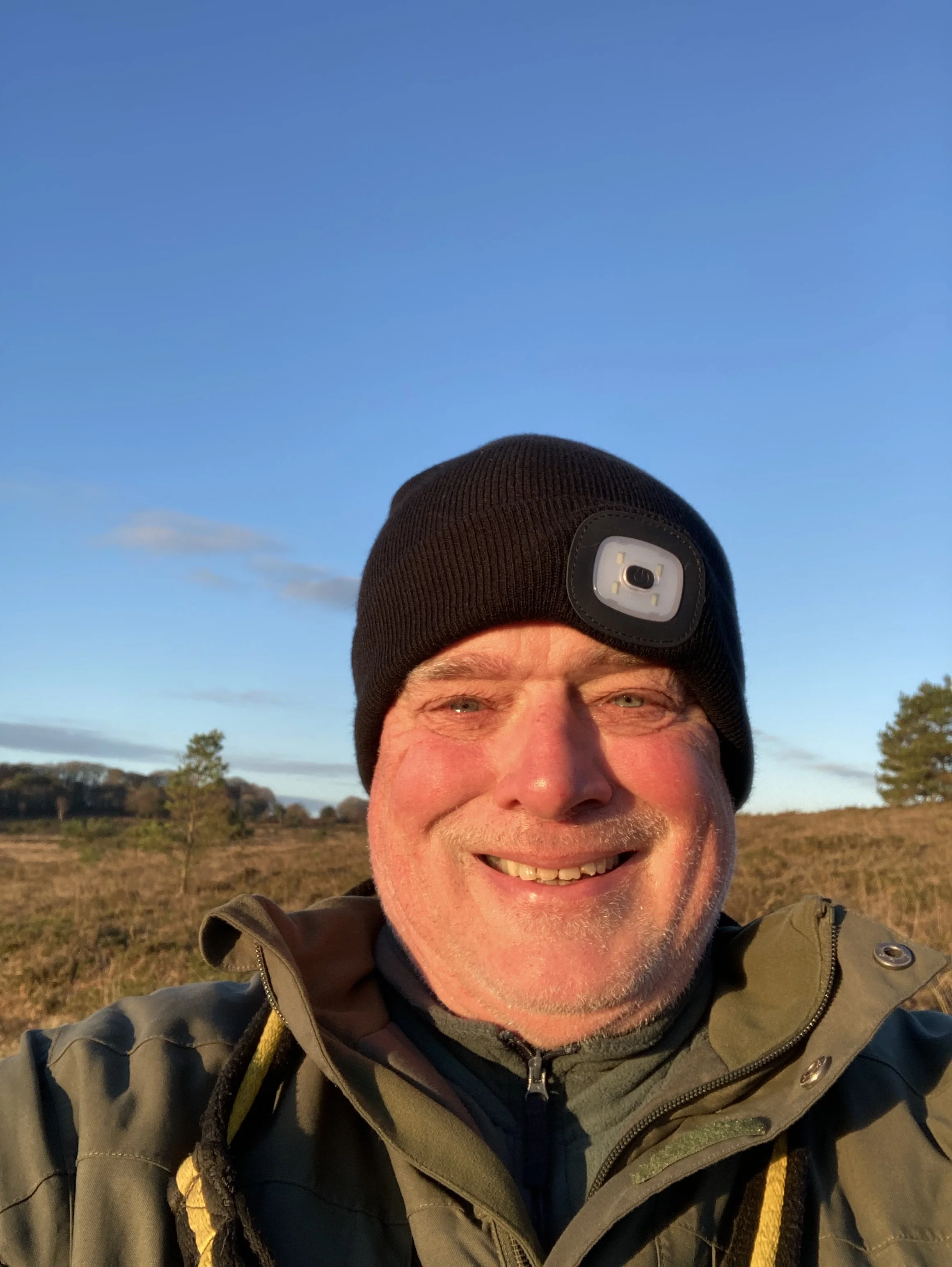 A smiling man wearing a black beanie with a headlamp, dressed in outdoor clothing, standing in a grassy field during sunset with a clear blue sky and trees in the background.