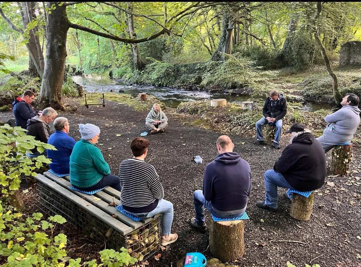 A group of people sits in a circle in a wooded area by a creek, participating in an outdoor meeting or gathering. They are seated on benches and tree stumps, with some looking toward a woman sitting cross-legged in the center.