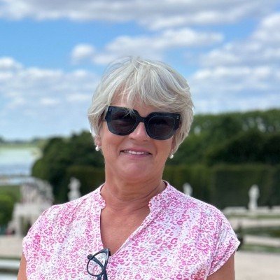 Smiling elderly woman with short white hair, wearing sunglasses and a pink patterned shirt, outdoors with trees and a cloudy sky in the background.
