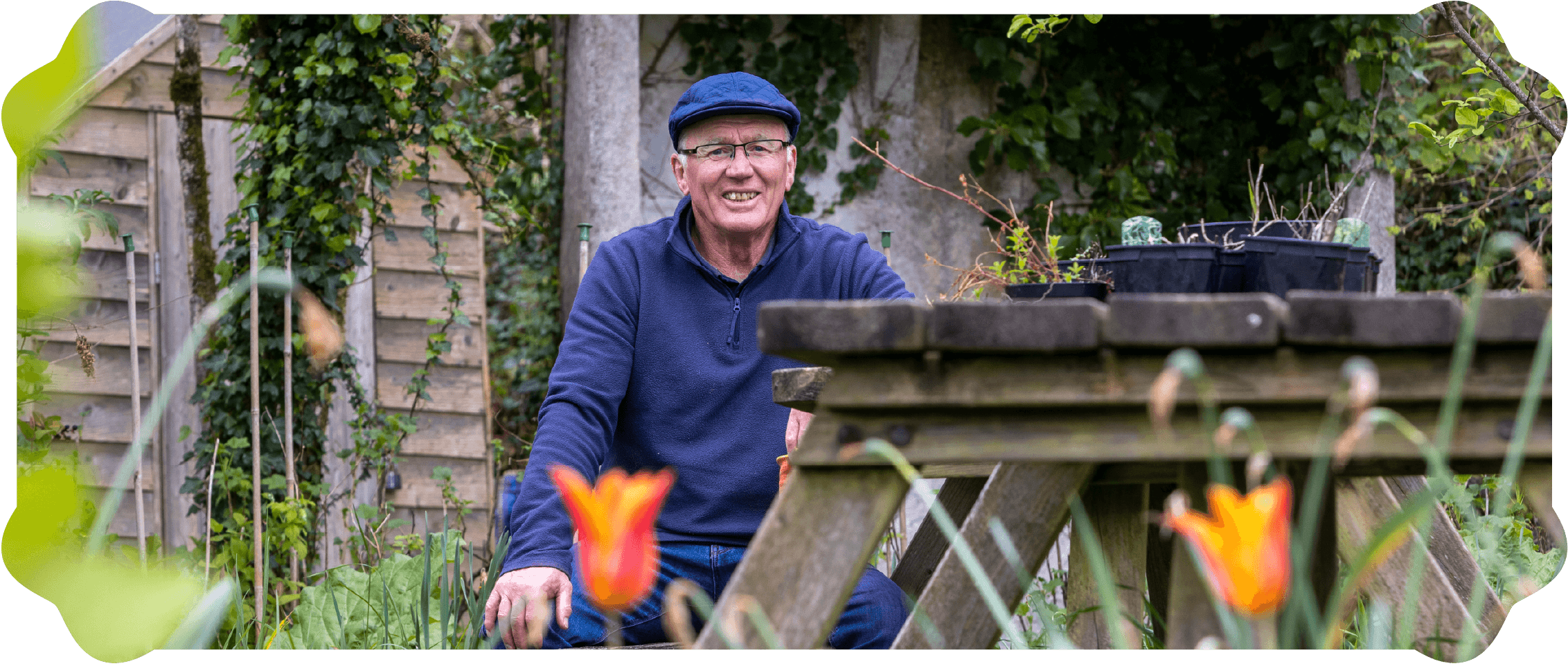 An elderly man wearing glasses, a blue cap, and a dark blue jacket, smiling while sitting in a garden surrounded by plants, flowers, and gardening tools.