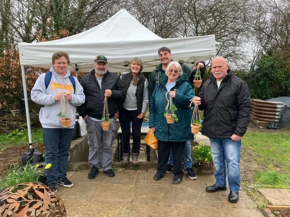 Six people standing outdoors in front of a white canopy tent, holding small hanging planters with greenery. The group appears cheerful, with some smiling and looking at the camera. The background features trees and garden elements.