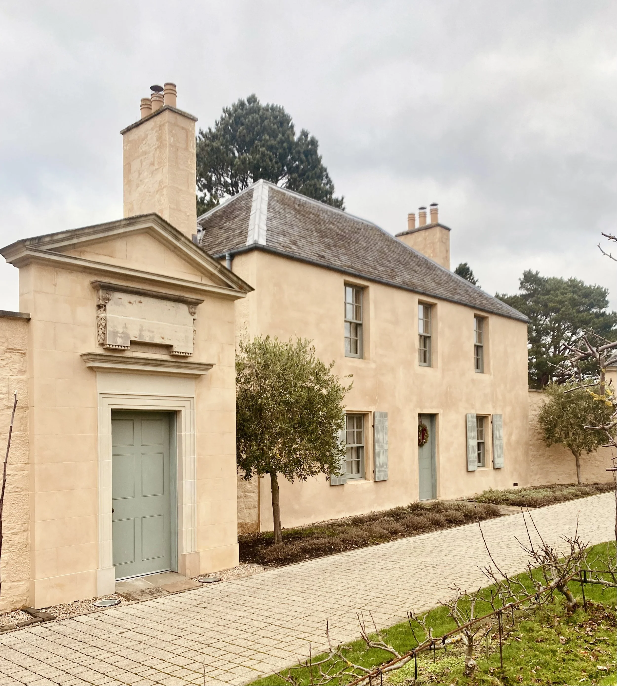 A historic beige house with blue shutters and multiple chimneys, situated alongside a cobblestone walkway, with small trees and a garden in the front.