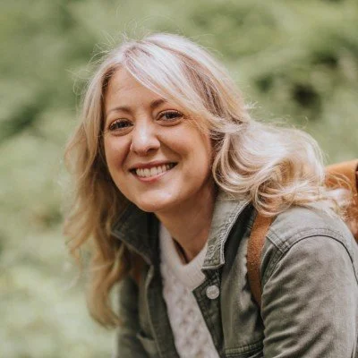 A smiling woman with blonde hair, wearing a gray jacket, outdoors in a green, blurred background