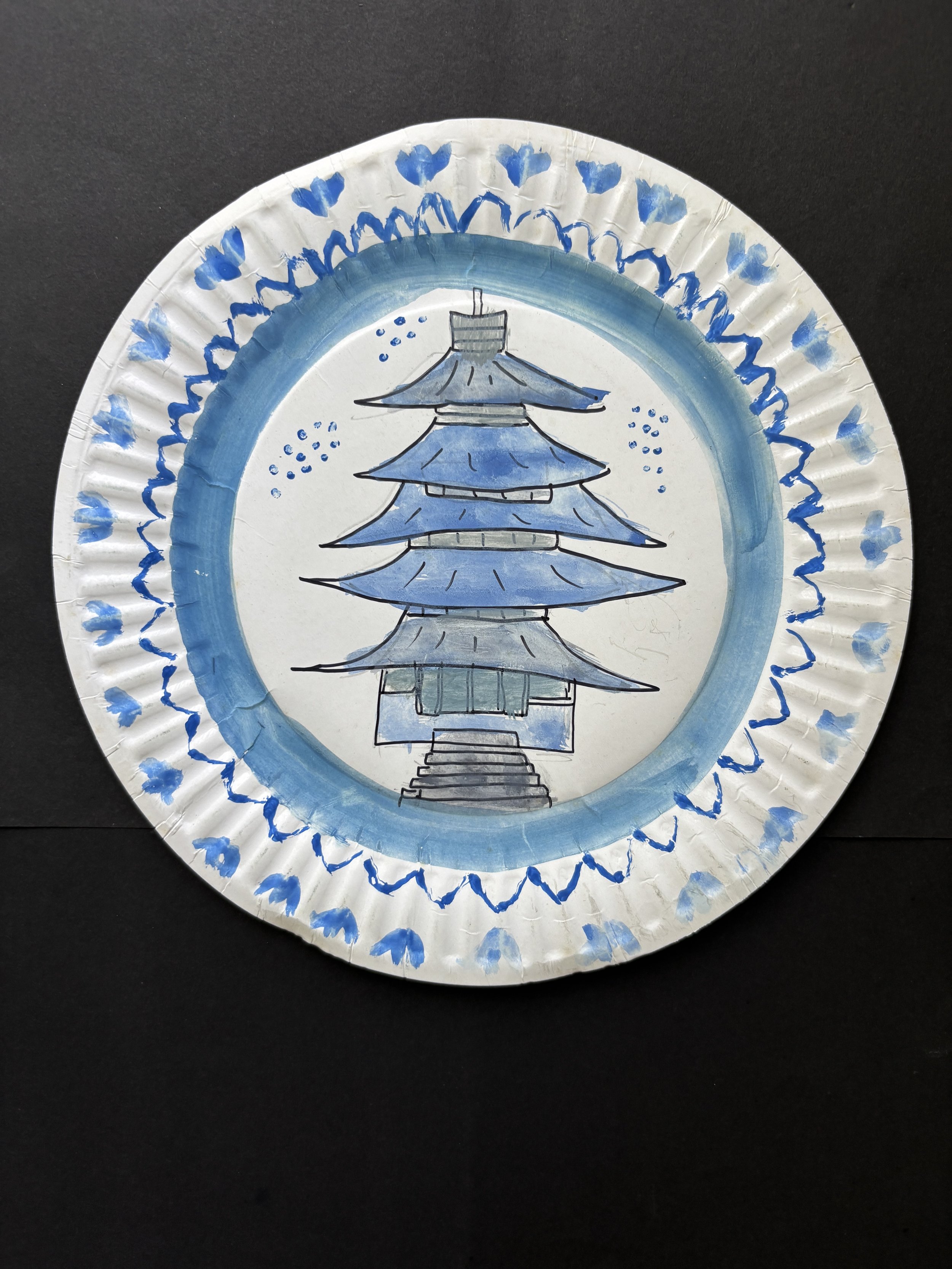 Decorative paper plate with a hand-painted blue and gray pagoda in the center, surrounded by a blue border and blue hearts along the edge.