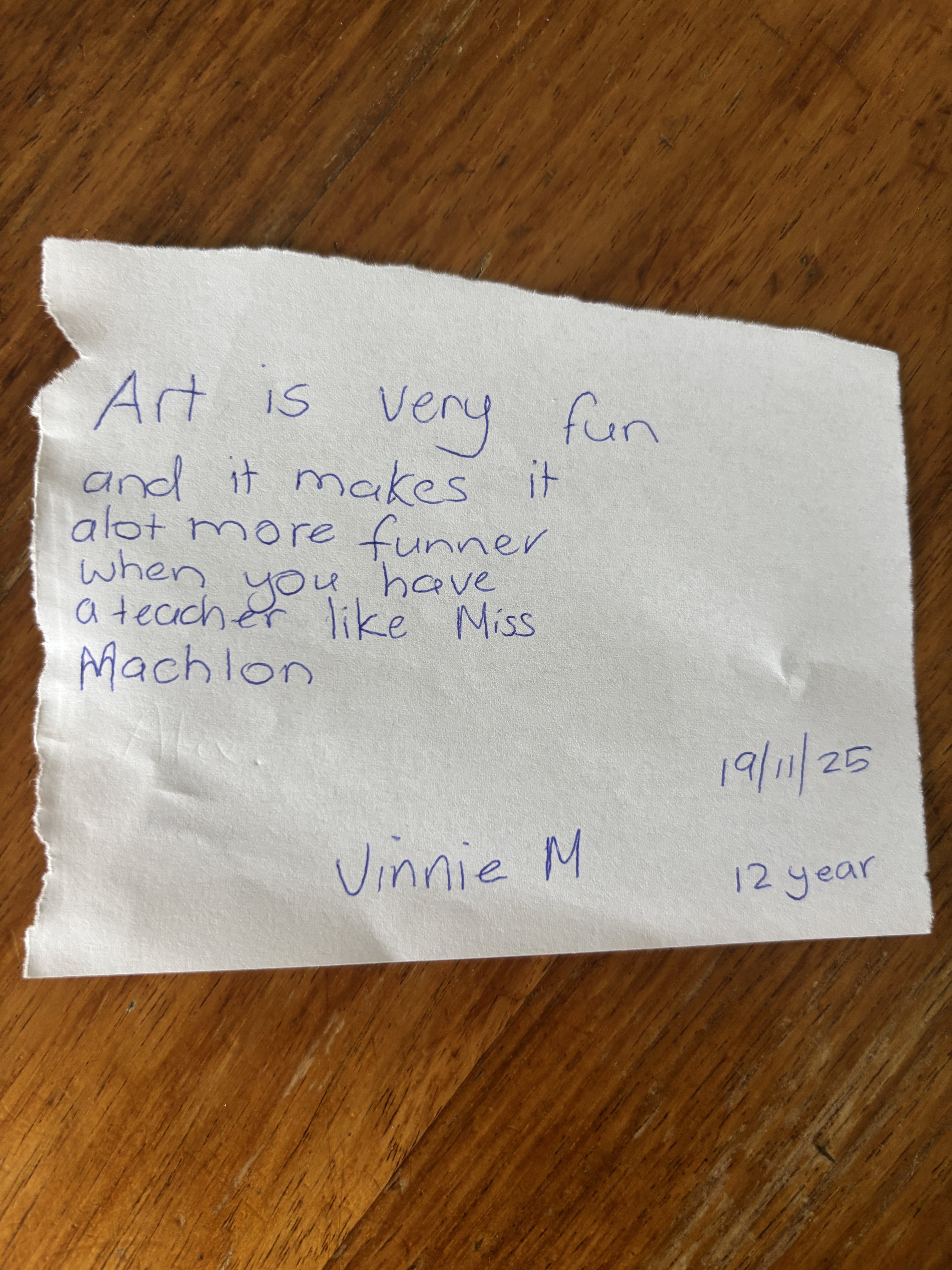 A torn piece of paper with handwritten note in blue ink, stating "Art is very fun and it makes it alot more funner when you have a teacher like Miss Machon". The note is signed by Vinnie M, dated 19/11/25, and mentions age 12 years. The paper is plac