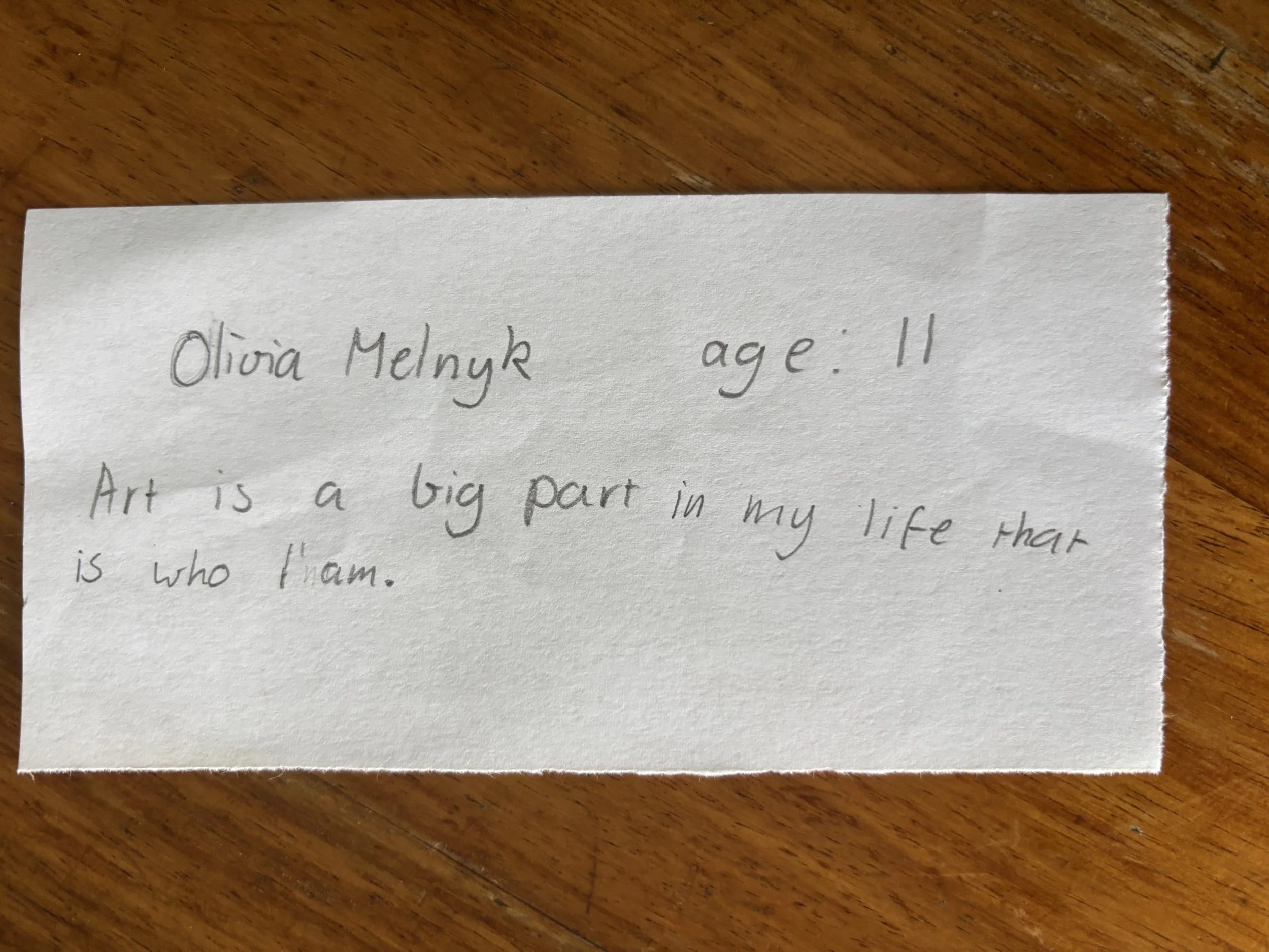 Handwritten note on a piece of paper placed on a wooden surface. The note contains personal details and a quote about art.