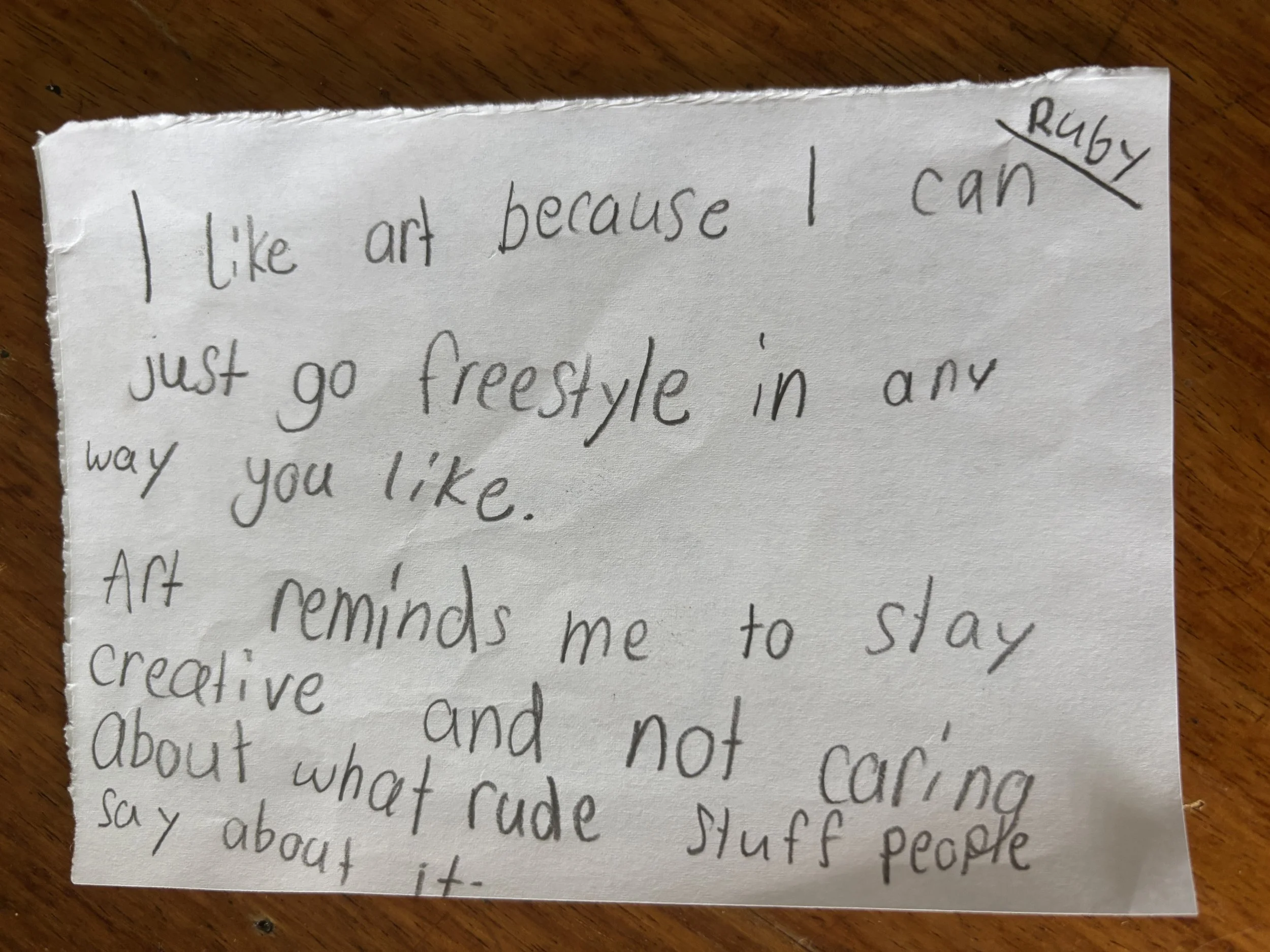 A handwritten note on a torn piece of paper that reads: "I like art because I can just go freestyle in any way you like. Art reminds me to stay creative and not caring about what rude stuff people say about it." The note has a small signature or name
