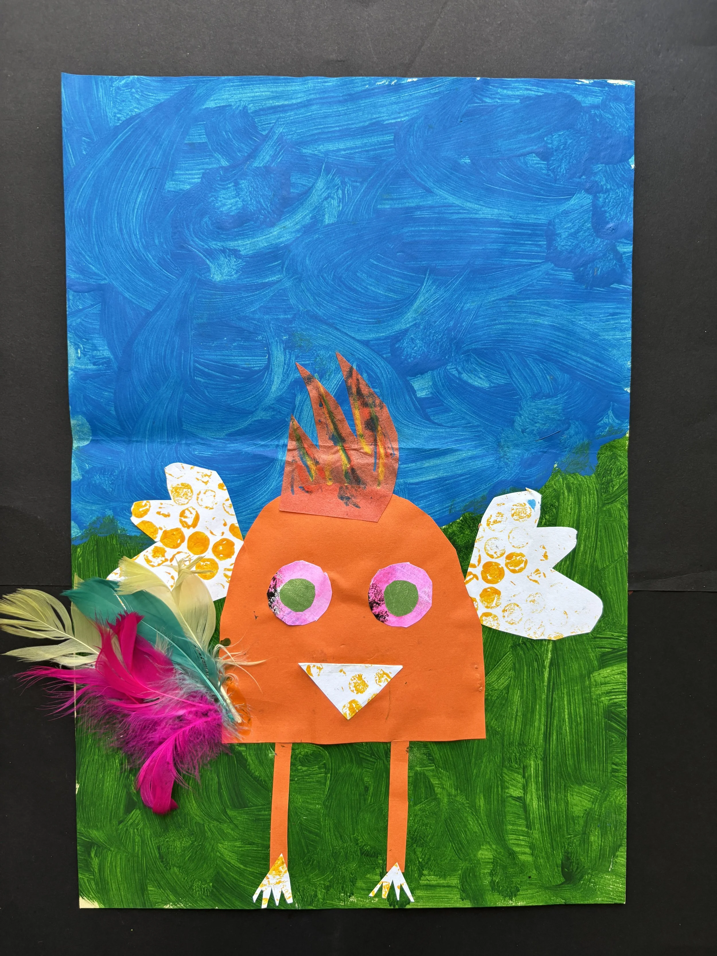 Colorful collage craft of a bird made from paper with feathers, set against a painted background of blue sky, green grass, and white clouds.