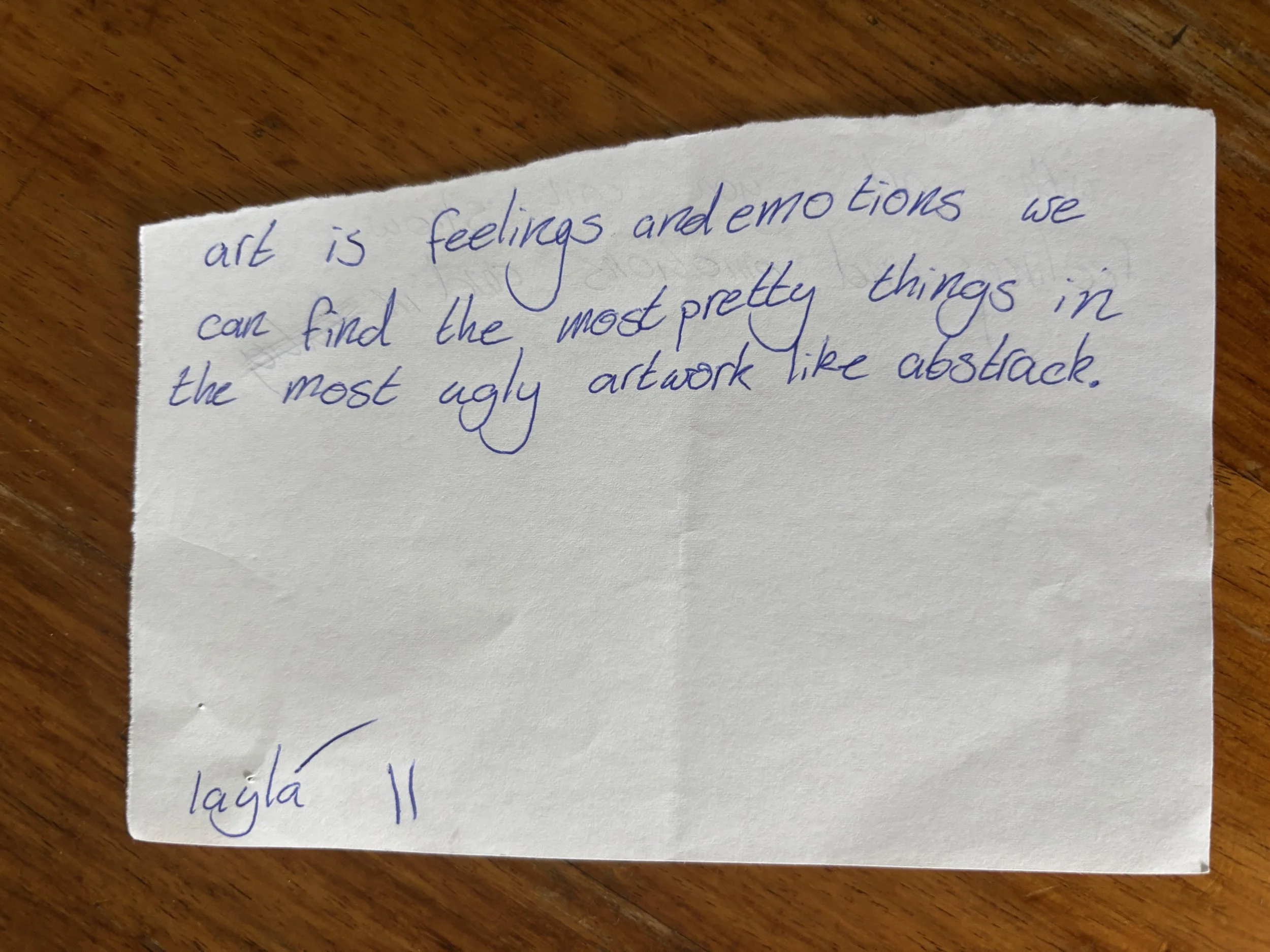 A handwritten note on a piece of torn white paper with blue ink, resting on a wooden surface. The note reads: "art is feelings and emotions we can find the most pretty things in the most ugly artwork like abstract." The bottom left corner of the pape