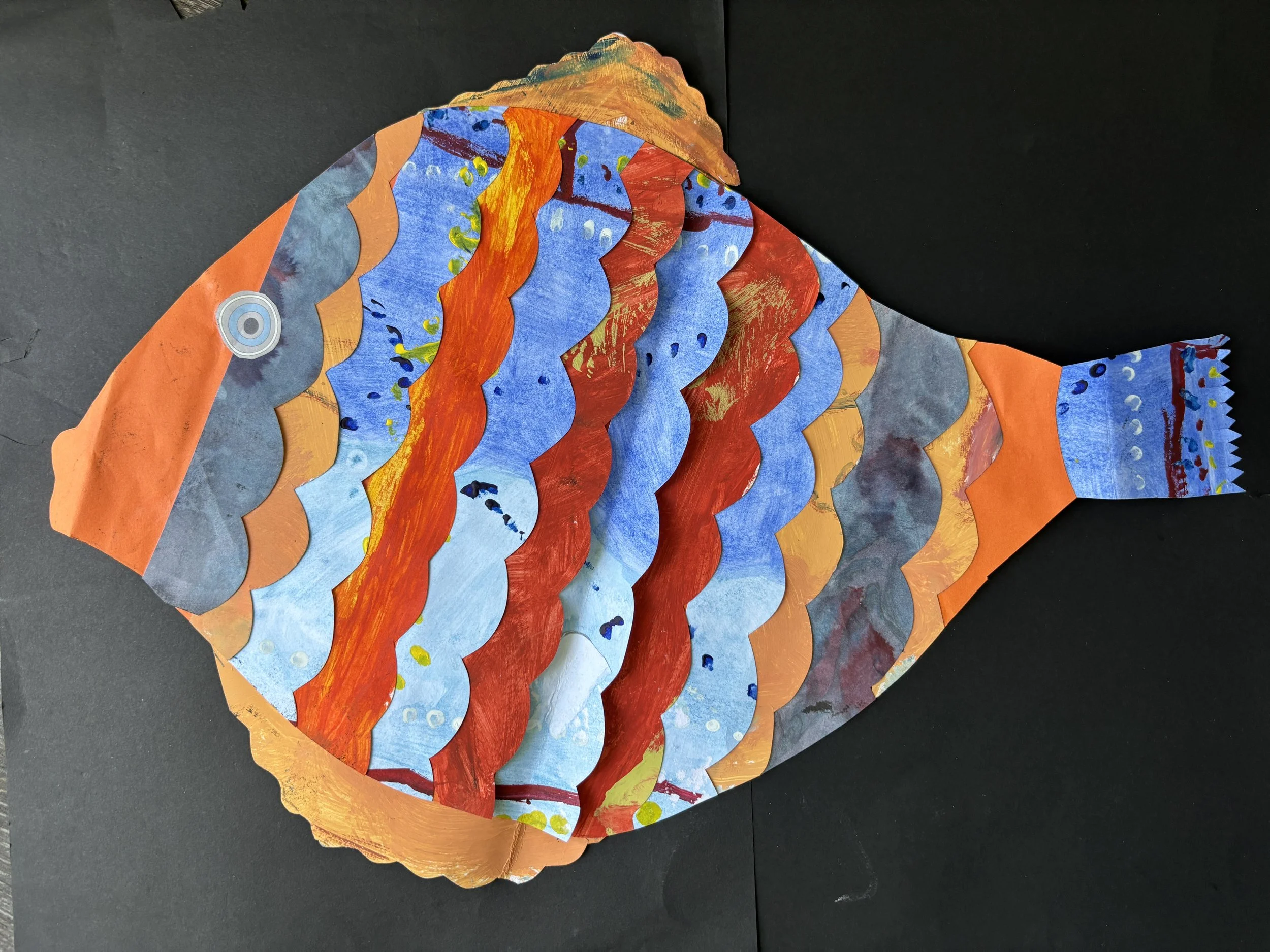 Colorful paper fish craft with layered fish scales in shades of orange, blue, red, and gray, on a black background.