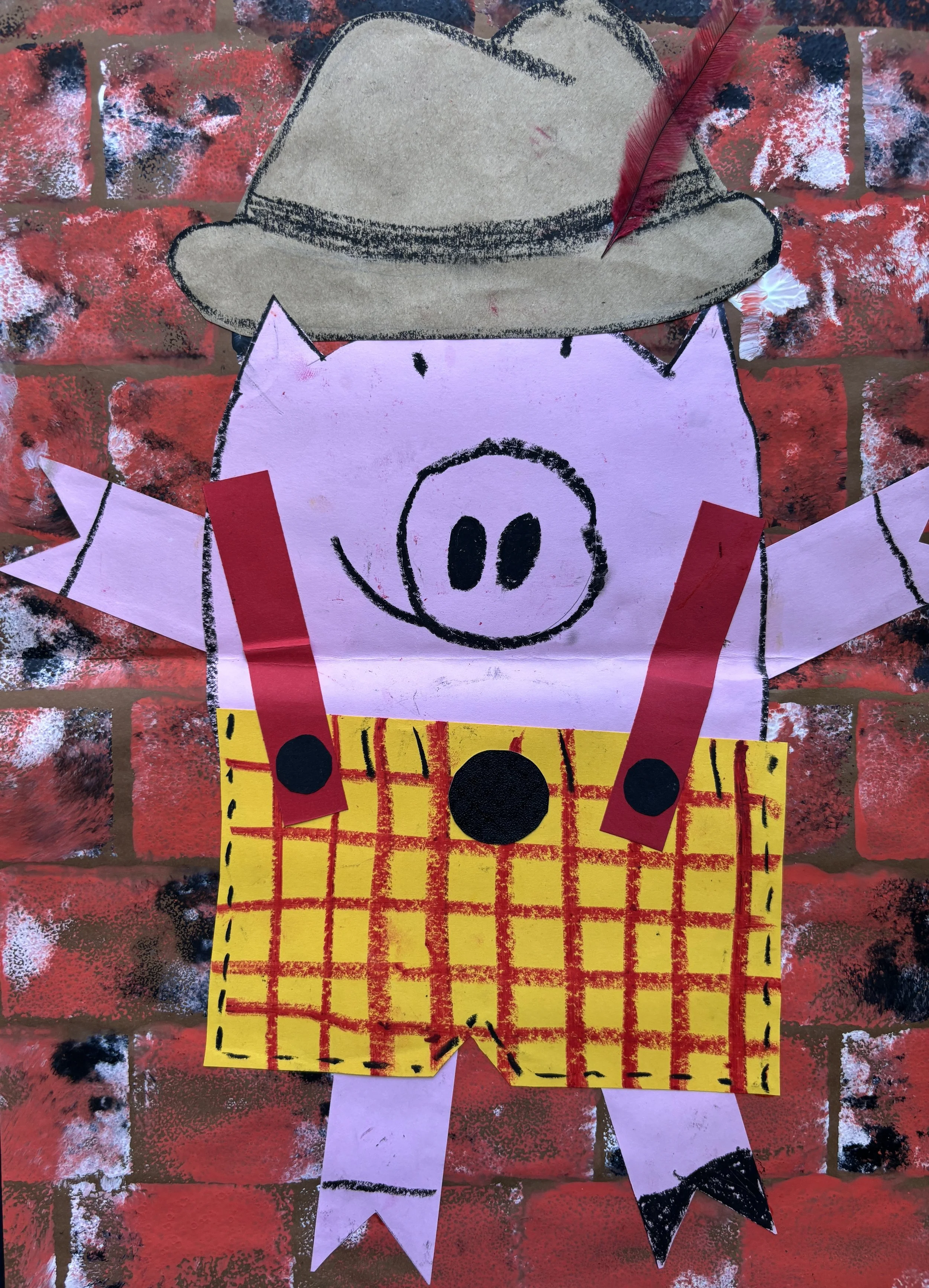 A colorful pig craft with a pink body, black eyes, and snout, wearing a beige hat with a red feather, a yellow plaid shirt, and red suspenders, mounted on a brick wall background