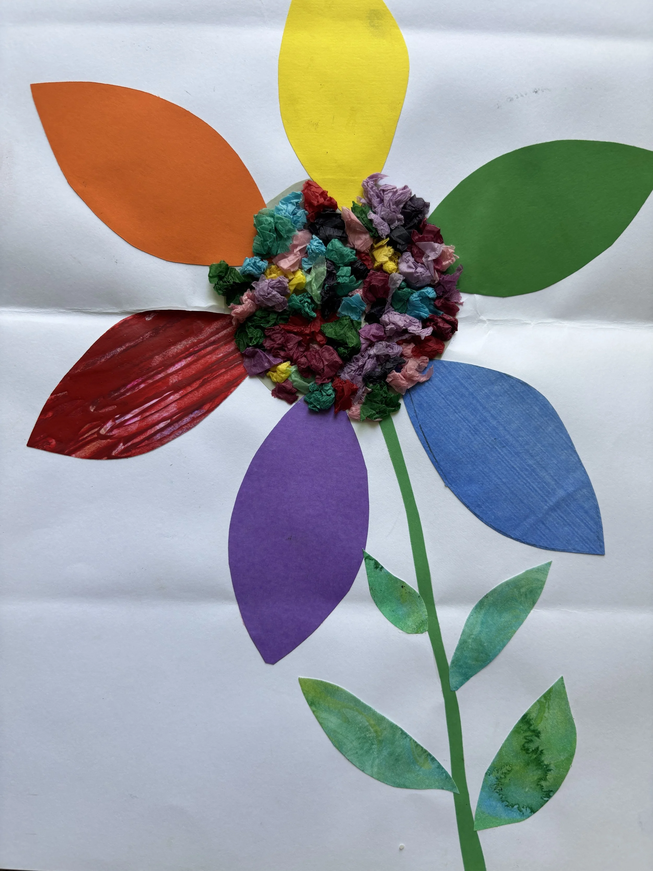 Colorful paper collage of a flower with six petals in orange, yellow, green, blue, purple, and red, with a center made of crumpled multicolored tissue paper and green paper leaves and stem.