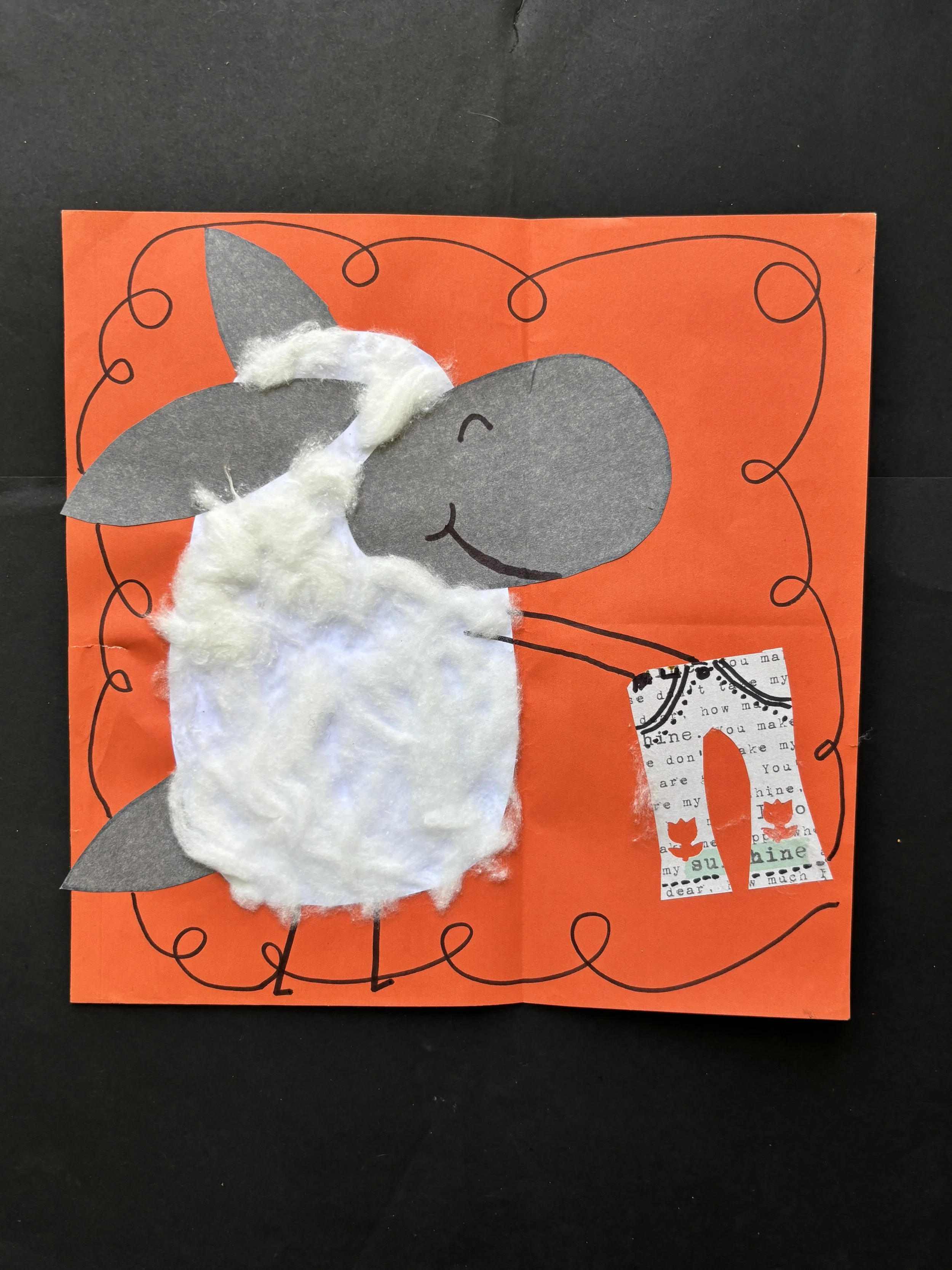 A handmade card with a cartoon sheep made from gray paper for the head and ears, white cotton for the body, and black lines for details, on an orange background with black squiggly border lines and the letter 'A' decorated with two small red cats.