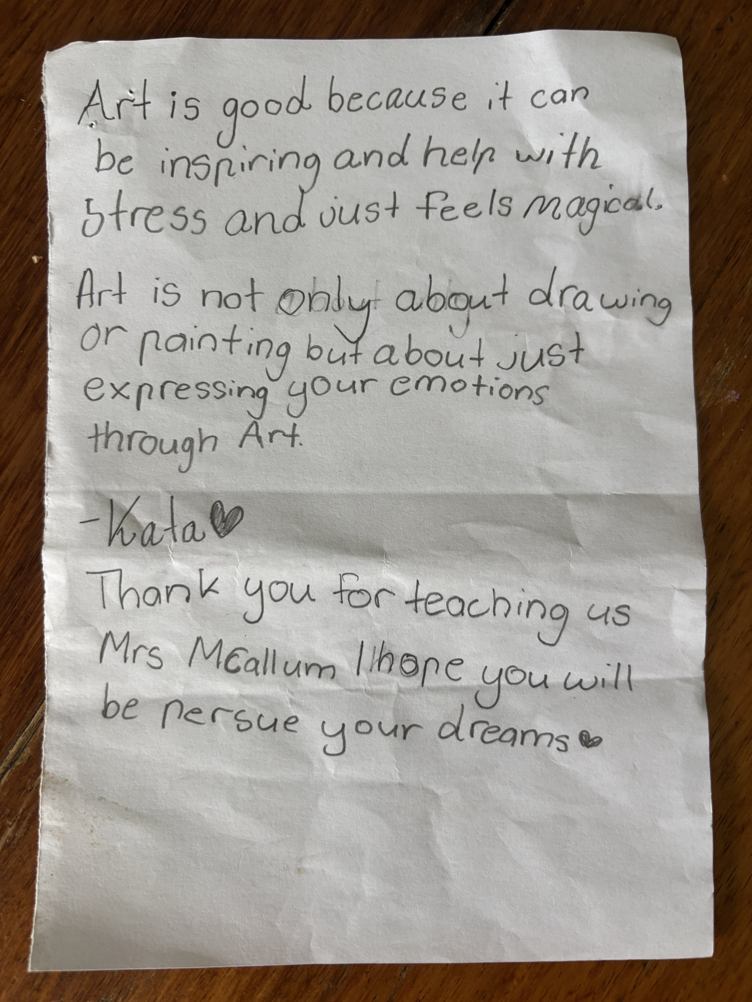 A handwritten note on a crumpled piece of white paper with black ink. The note discusses the importance of art, expresses gratitude to a teacher, and wishes her success in pursuing her dreams. The note is signed by 'Katal' with two small hearts next 