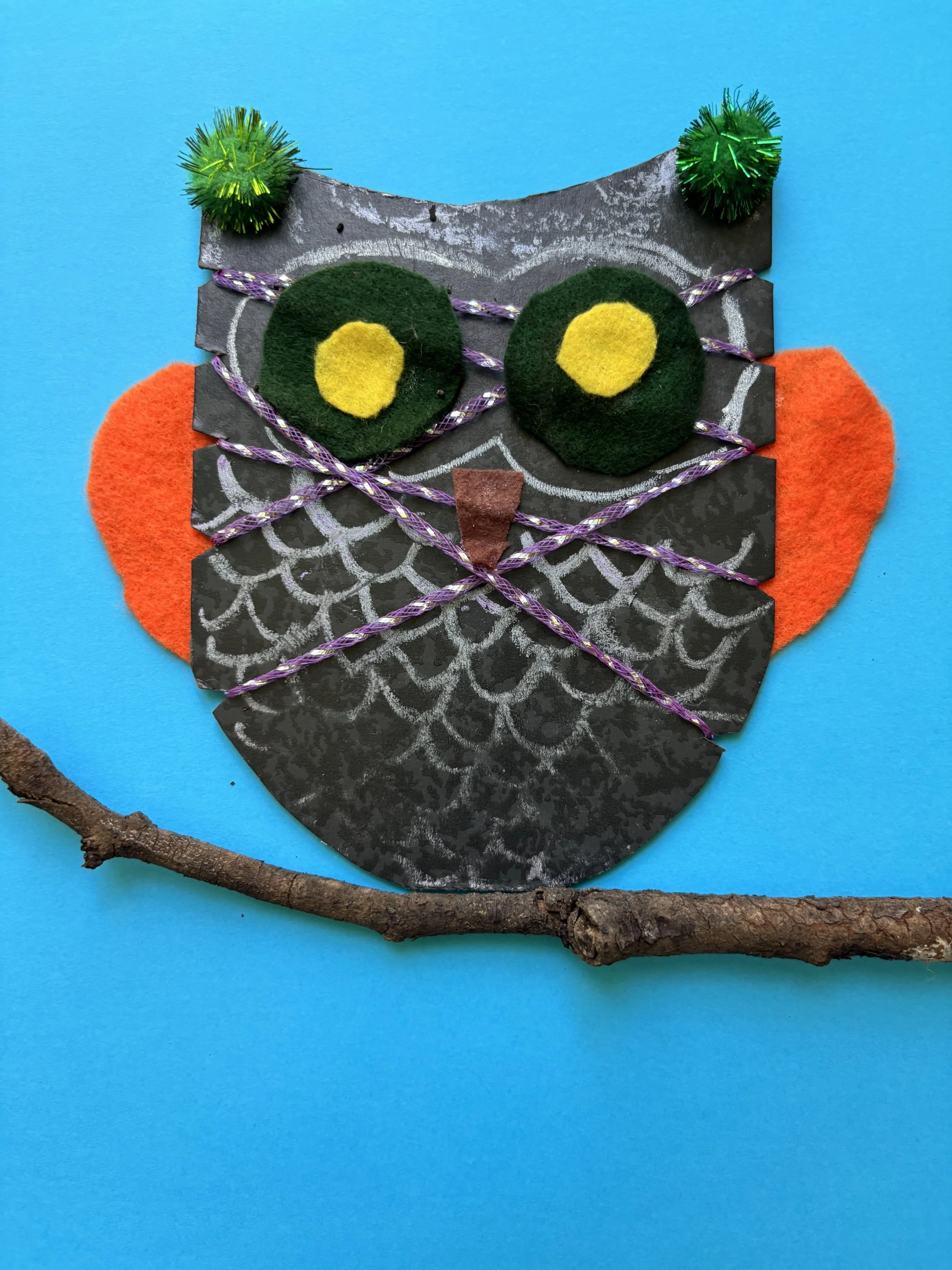Craft owl made of felt, paper, and string on a blue background, with a small branch at the bottom.