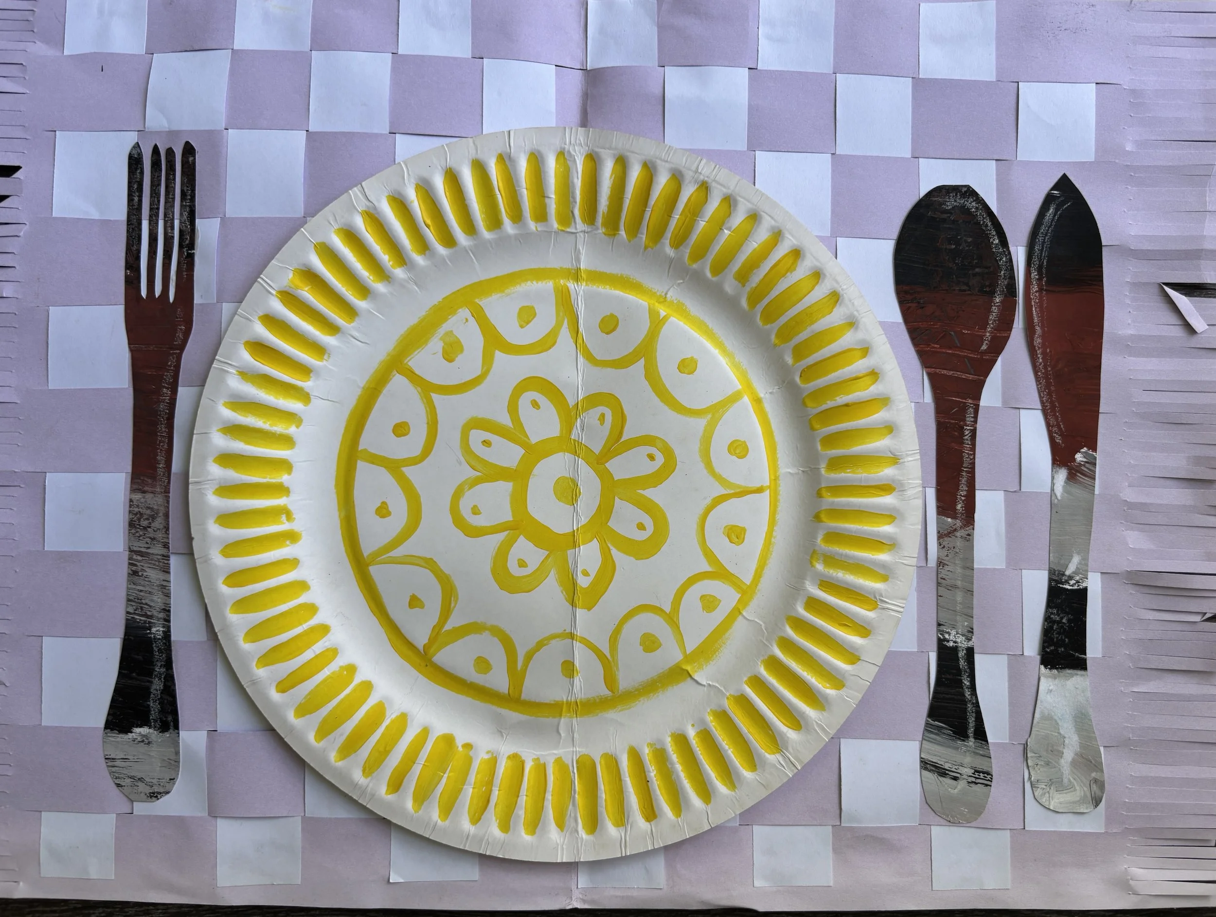 Decorative paper plate with a yellow floral and geometric pattern, set on a pink and white checkered paper placemat. Flatware, a fork, a knife, and a spoon, are arranged on either side of the plate.