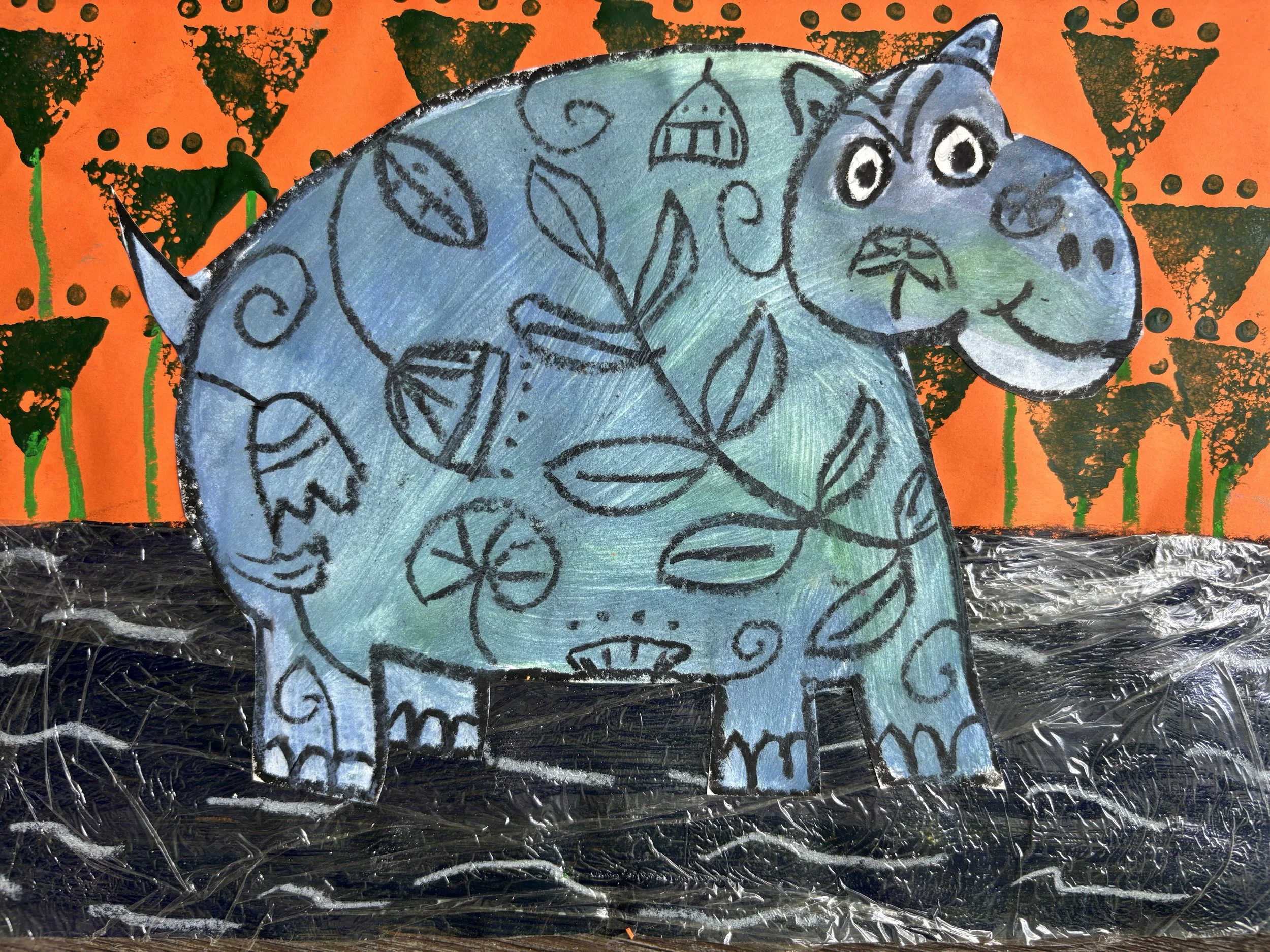 Children's artwork of a blue dinosaur with leaf and flower patterns on its body, set against an orange background with black triangles and green ribbons, on a black surface with plastic wrap.