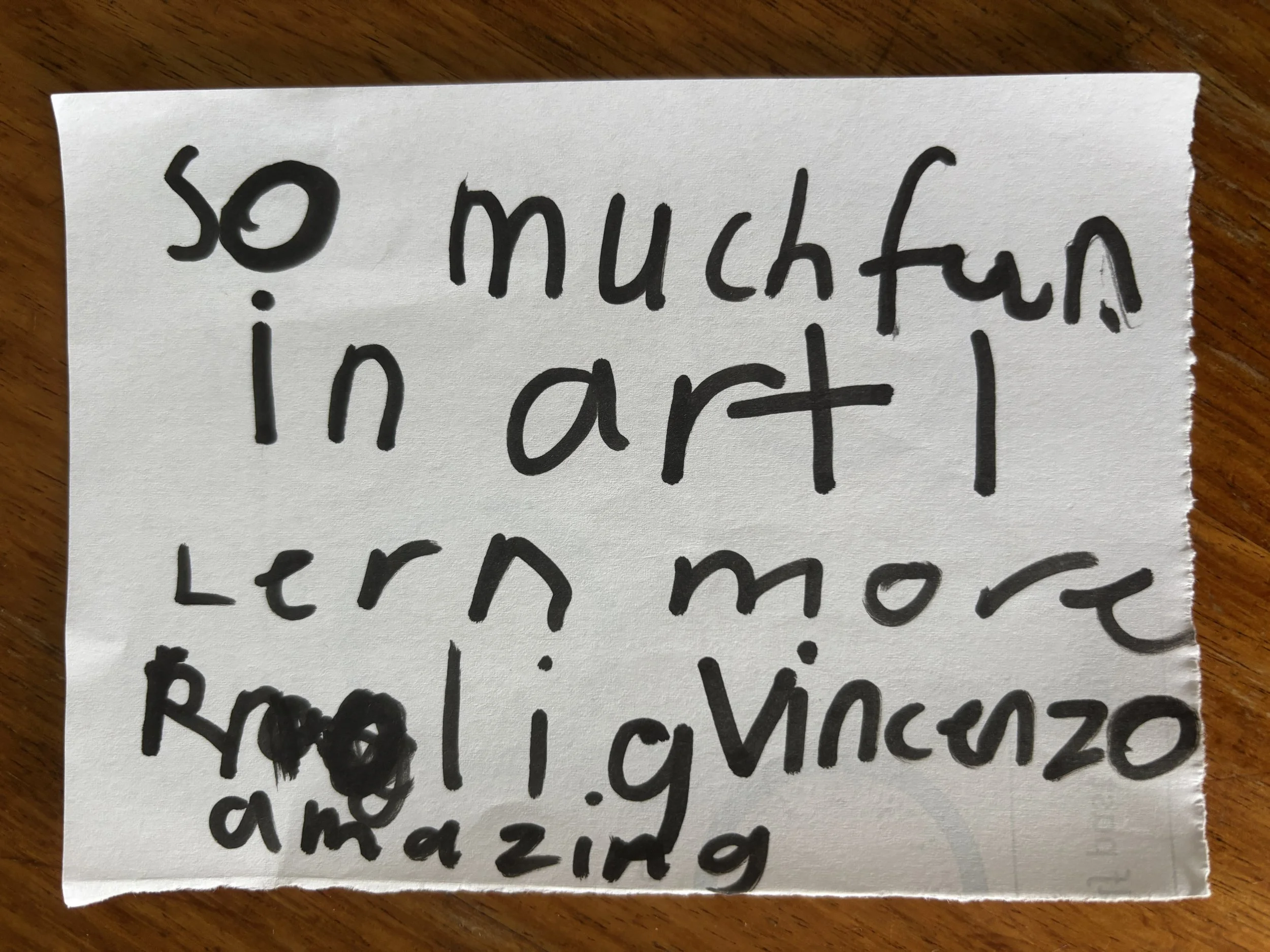 A handwritten note on a piece of white paper with black marker that says: 'so much fun in art I learn more, radi lvencio andazing'.
