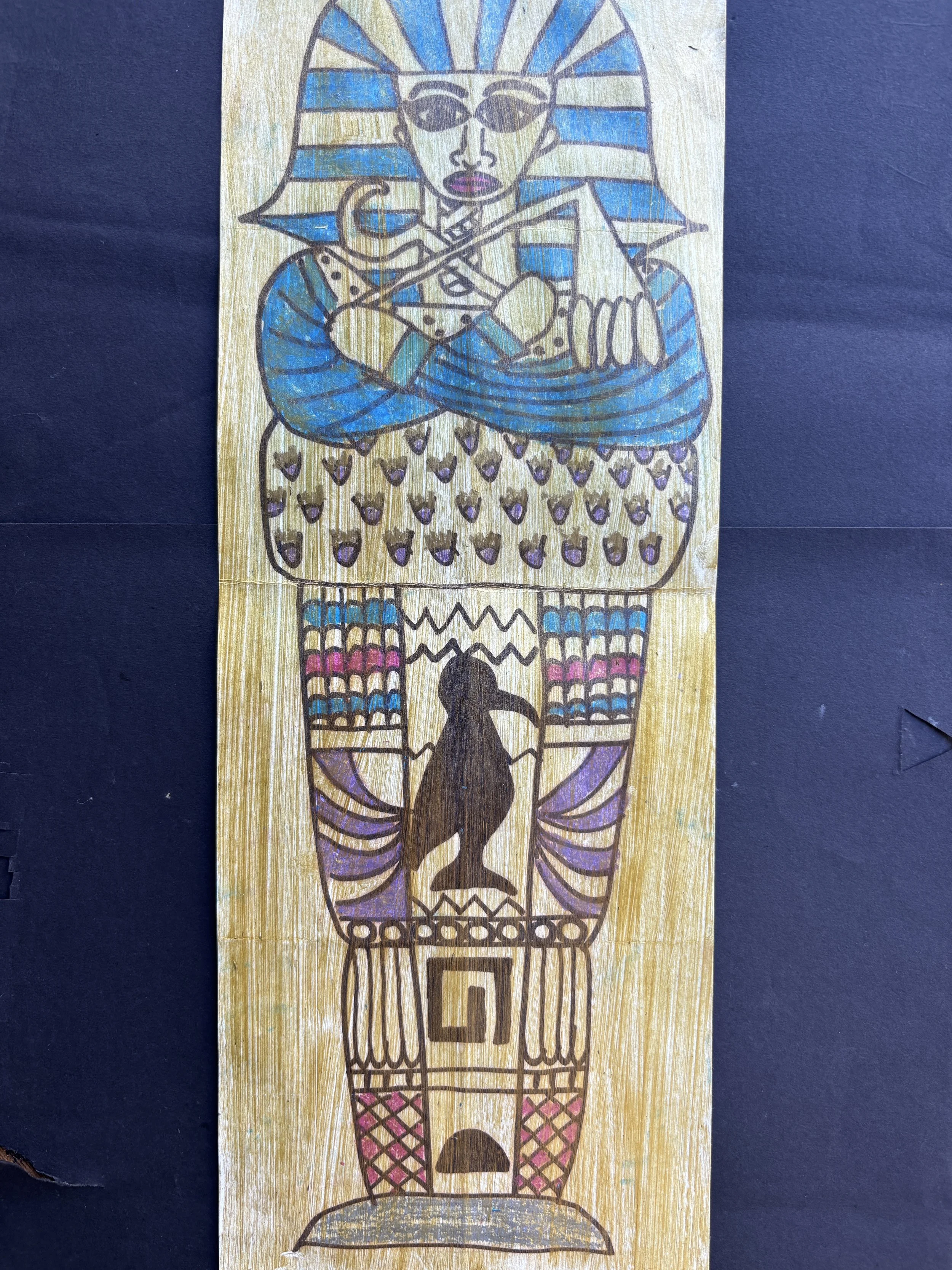 Colorful drawing of a woman with Egyptian headdress and sunglasses, holding a sword and a scepter, with a silhouette of a dog at the bottom, on a wooden background.