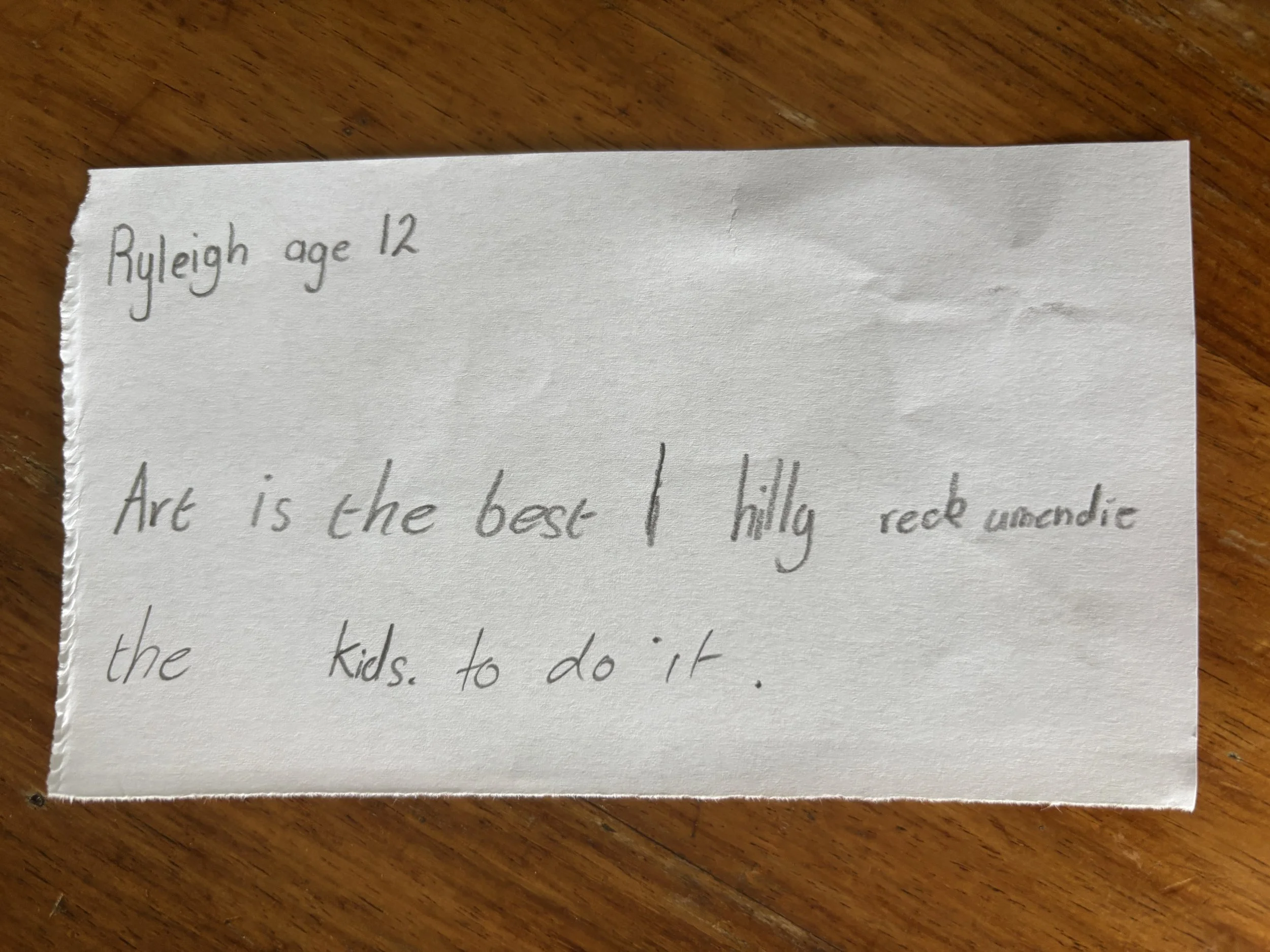 A handwritten note on a torn piece of paper with the text: "Ryleigh age 12. Art is the best I really think under the kids. to do it." The paper is placed on a wooden surface.