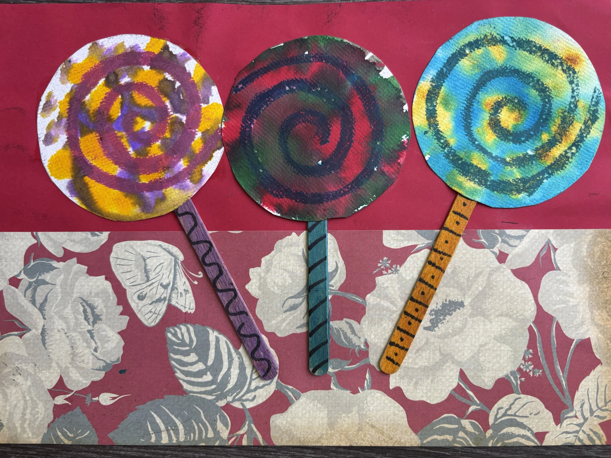 Three colorful, spiral-shaped paper crafts with painted handles on a pink and floral background.