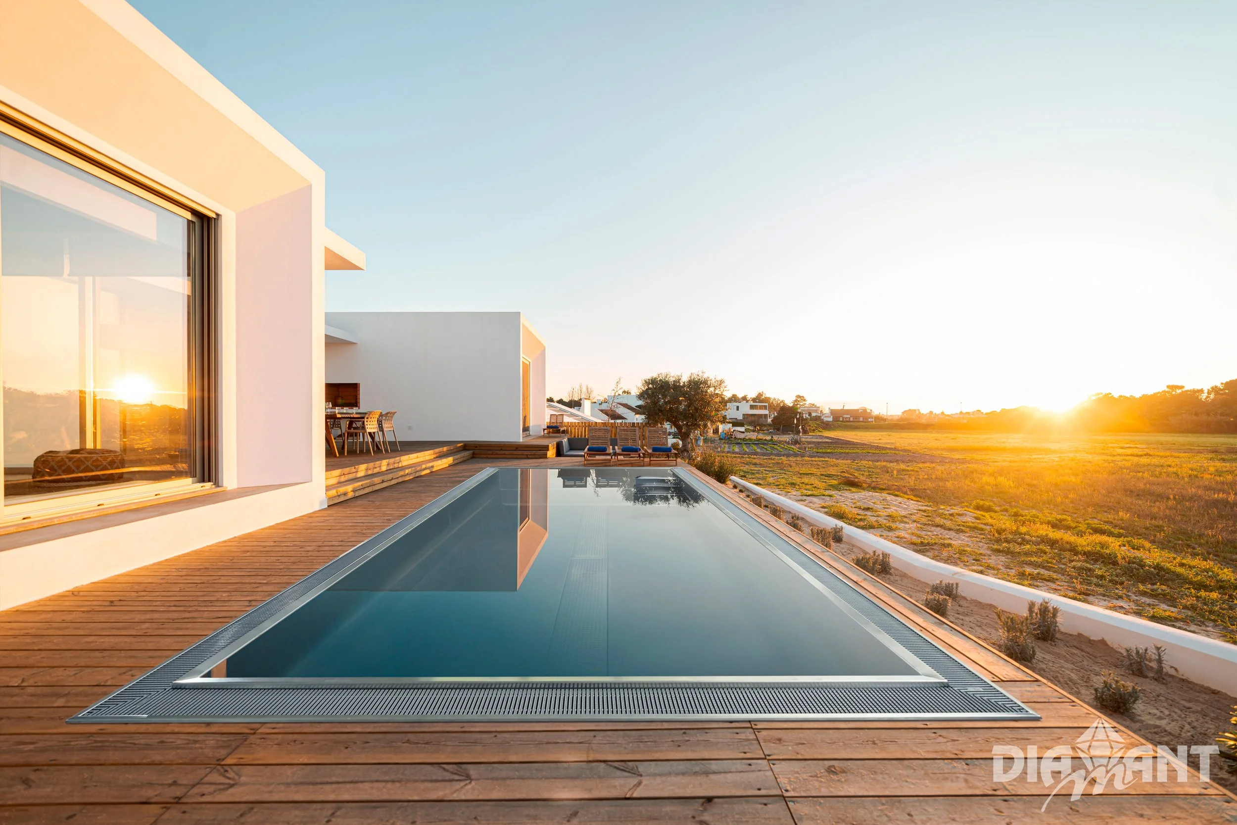 Modern house with a rectangular swimming pool on a wooden deck at sunset, overlooking open land with a tree and distant houses.