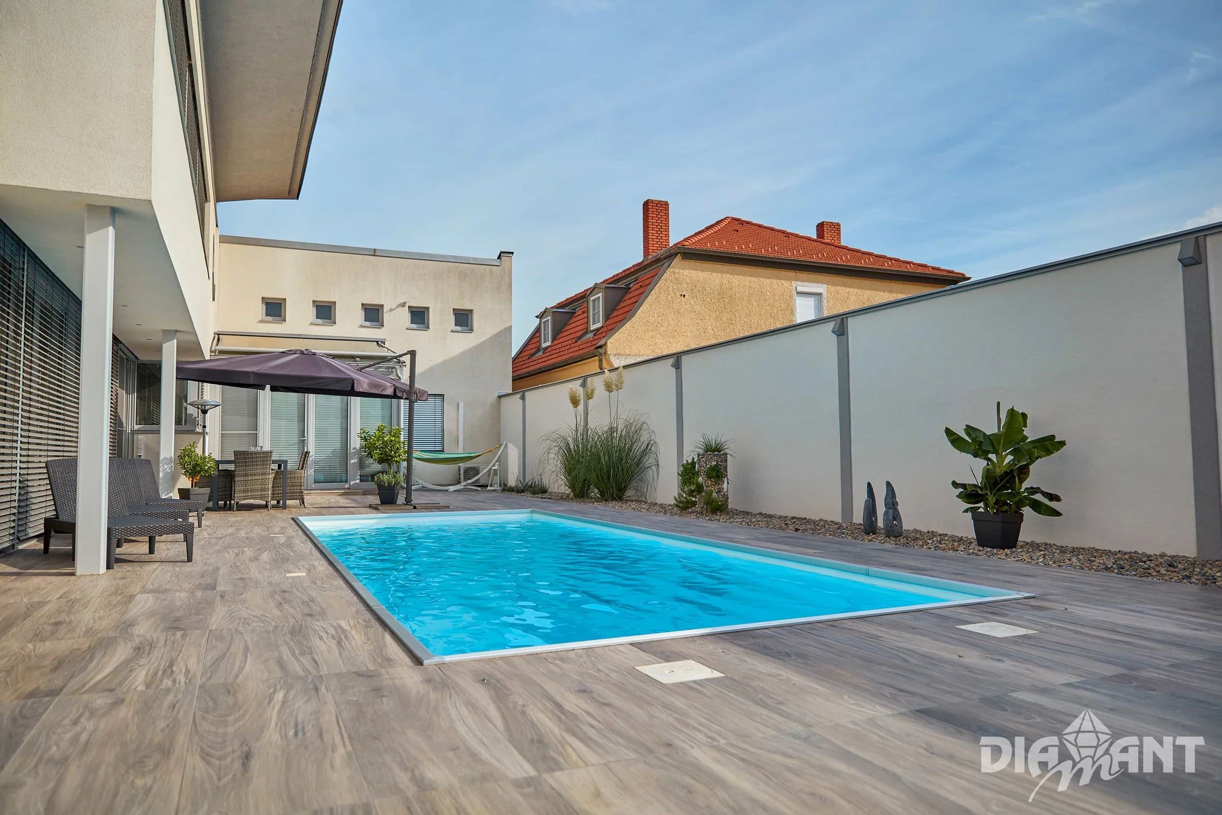 Empty backyard swimming pool with wooden deck, patio furniture including a table and chairs, an umbrella, a hammock, potted plants, and outdoor decor, enclosed by a white wall, with neighboring houses and a blue sky in the background.