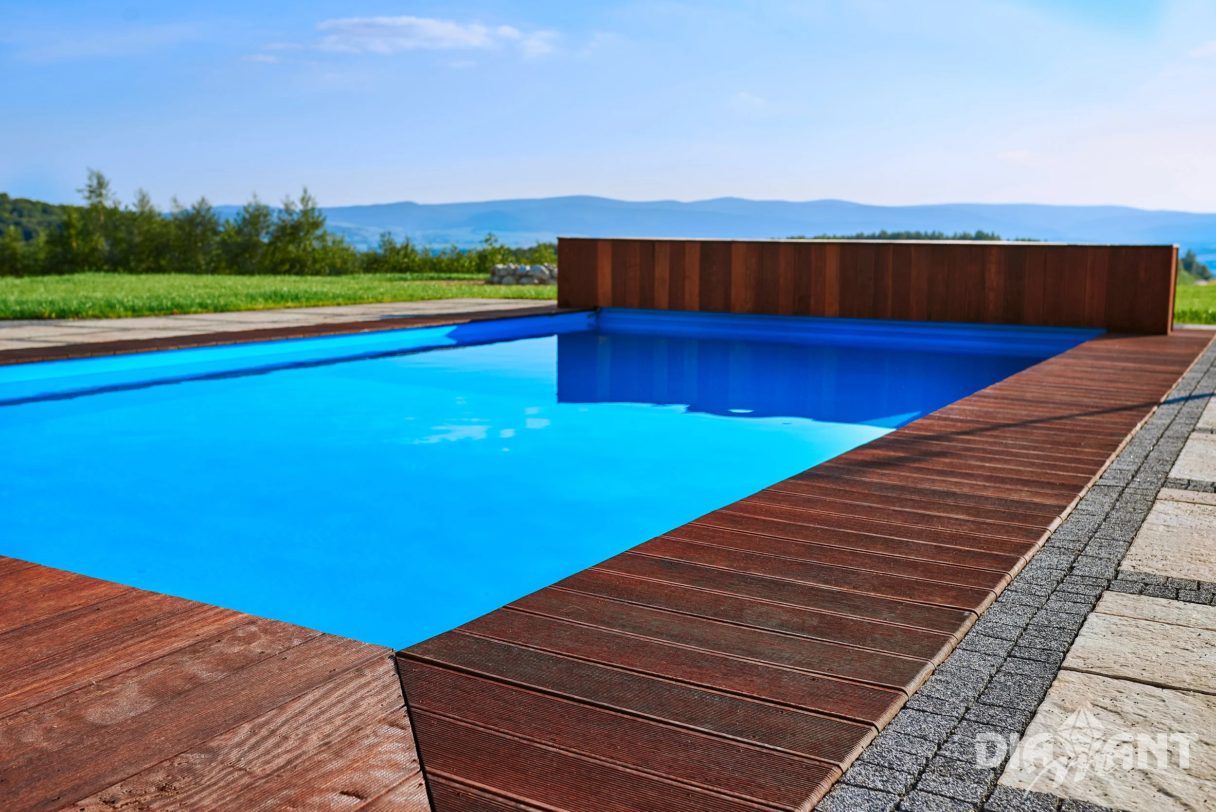 An outdoor swimming pool with bright blue water, surrounded by wooden decking and concrete pavers, with a scenic landscape and mountains in the background.