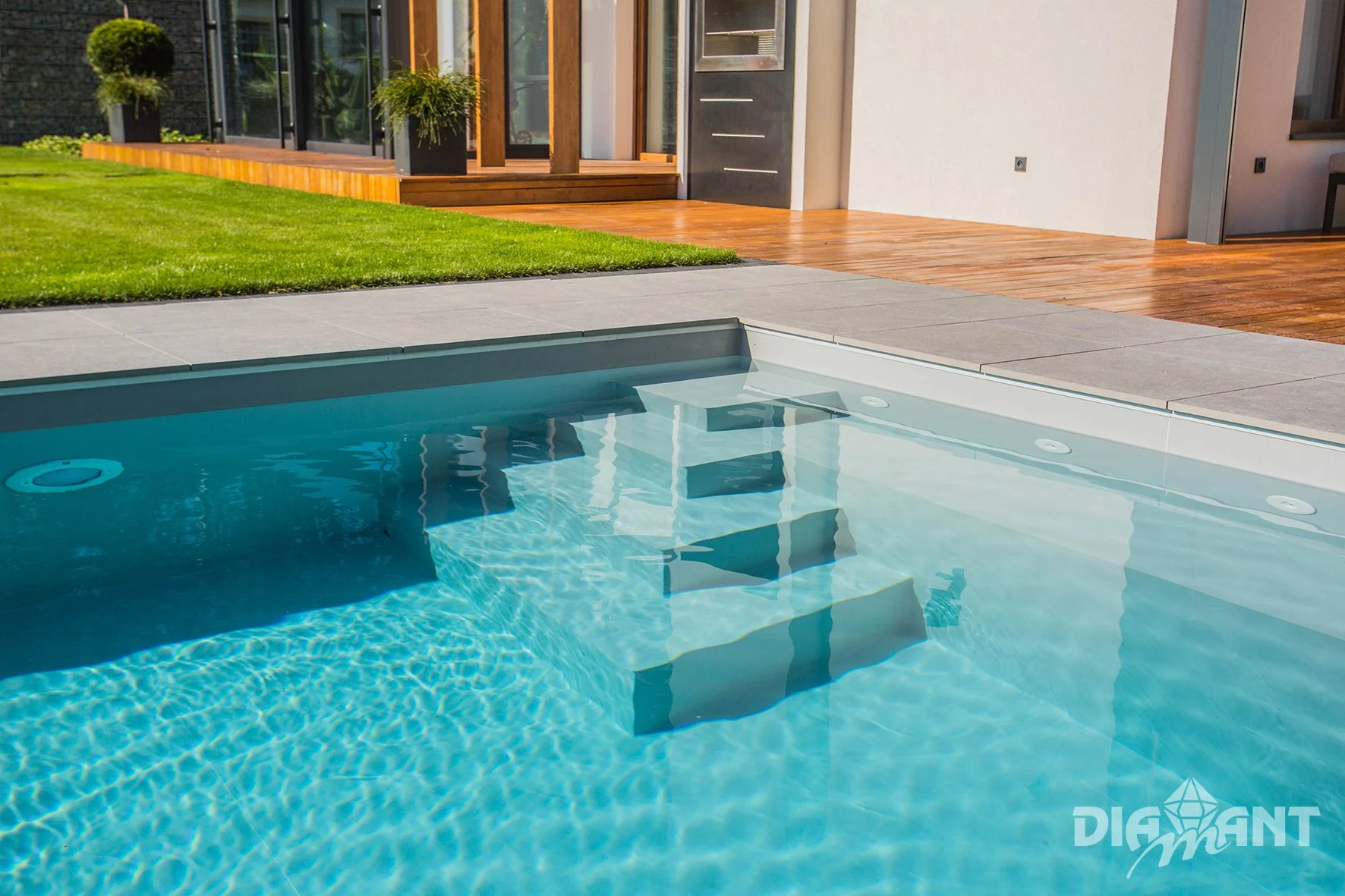 Modern backyard with a swimming pool, wooden deck, green lawn, and patio area adjacent to a contemporary house.