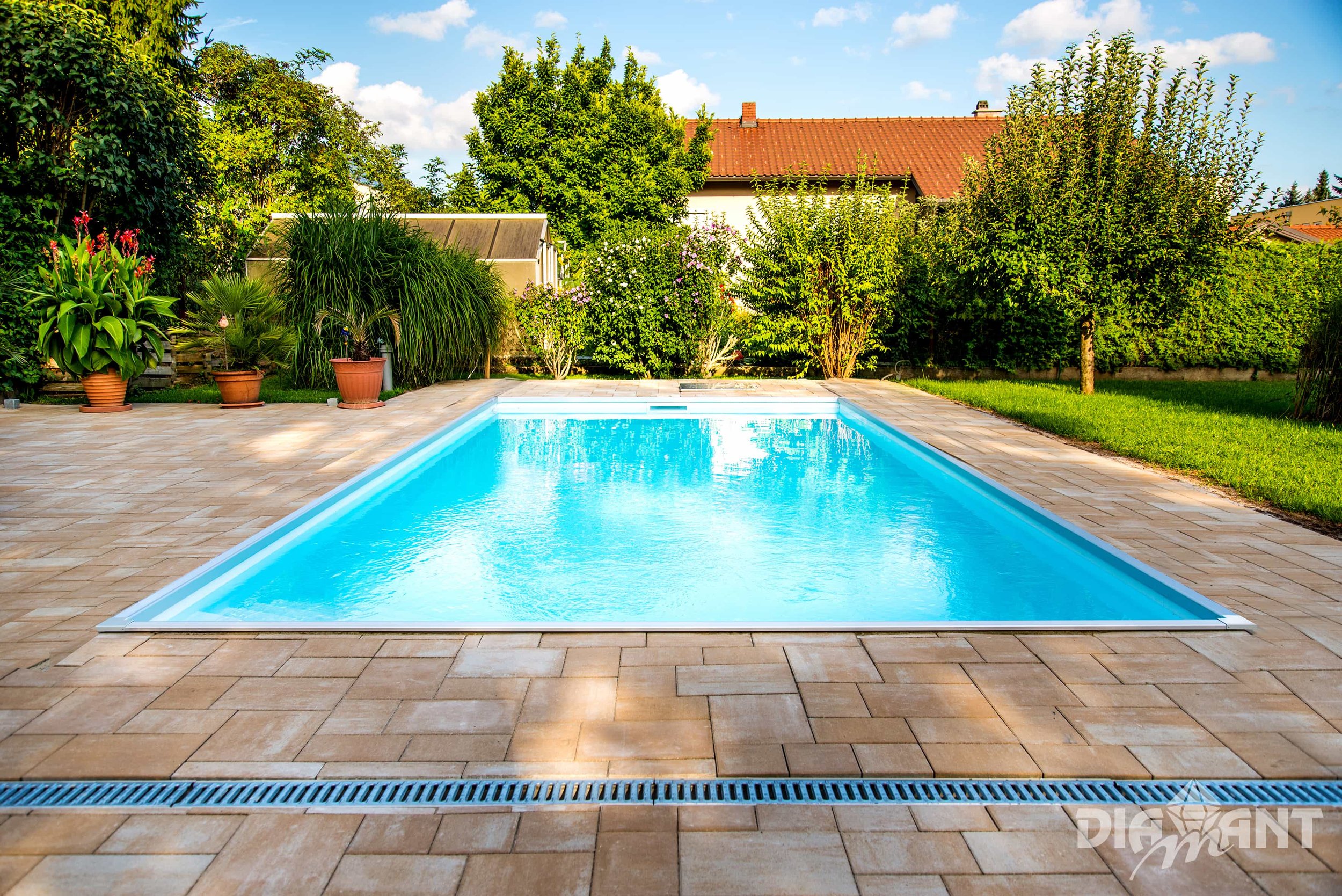 A backyard swimming pool surrounded by a tiled patio, lush green trees and shrubs, with a house with a red tile roof in the background on a sunny day.