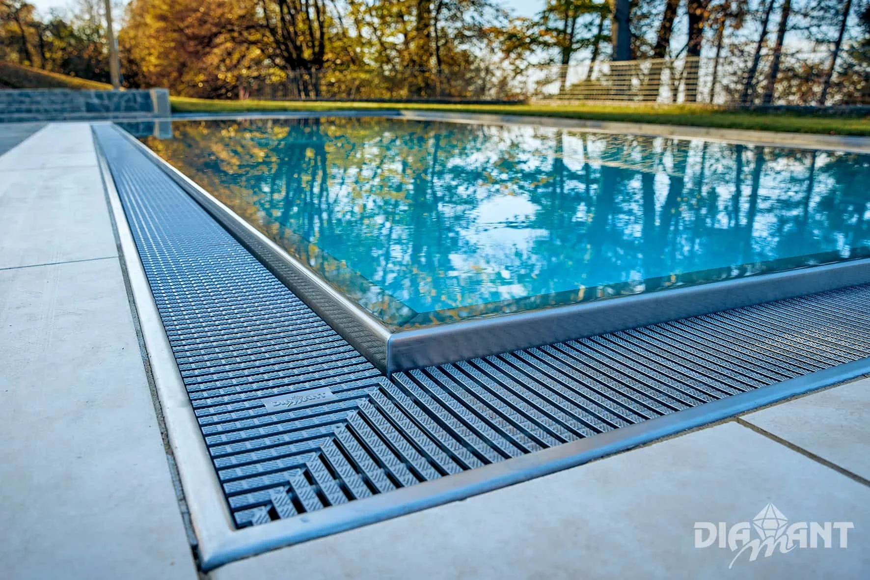 Close-up of the edge of an outdoor swimming pool with a metallic grate drain, with trees and a grassy area in the background during sunset.