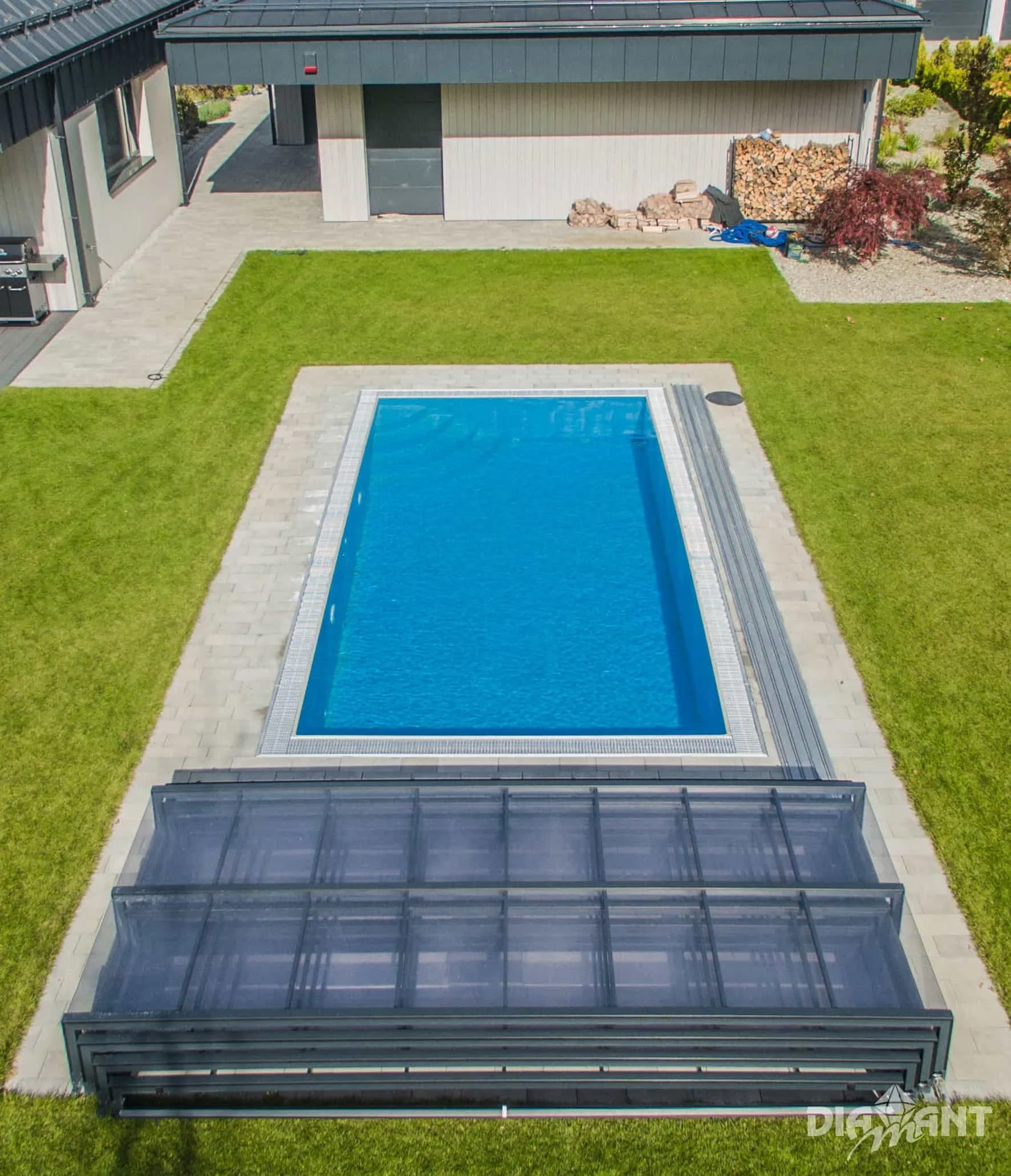 A backyard with a rectangular swimming pool, surrounded by a grassy lawn and a paved patio area. There is a covered structure with a transparent roof in the foreground, and a shed or garage along the back edge of the yard. The yard is landscaped with trees and bushes.