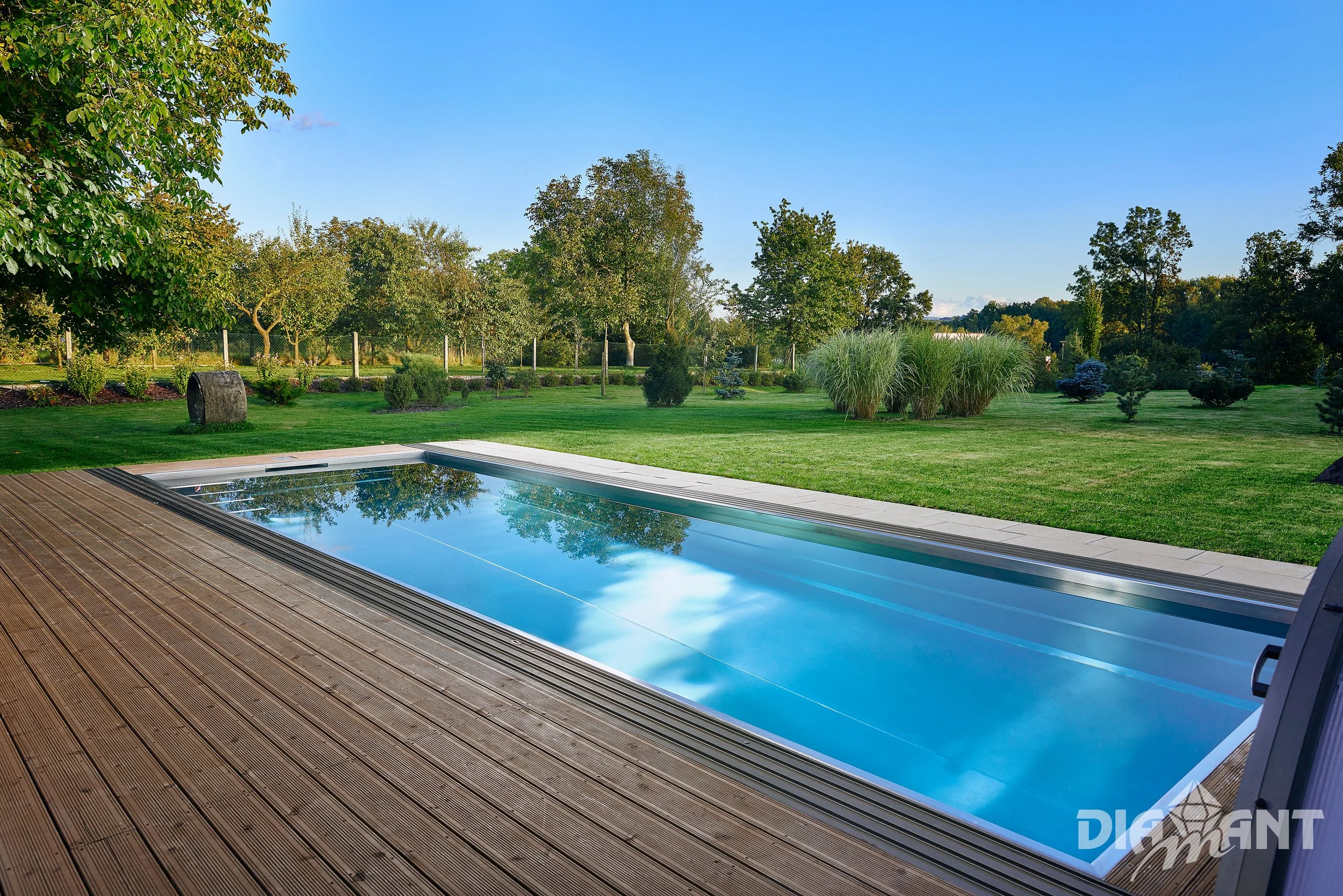 An outdoor scene featuring a covered rectangular swimming pool on a wooden deck, with a lush green lawn, trees, shrubs, and a clear blue sky in the background.