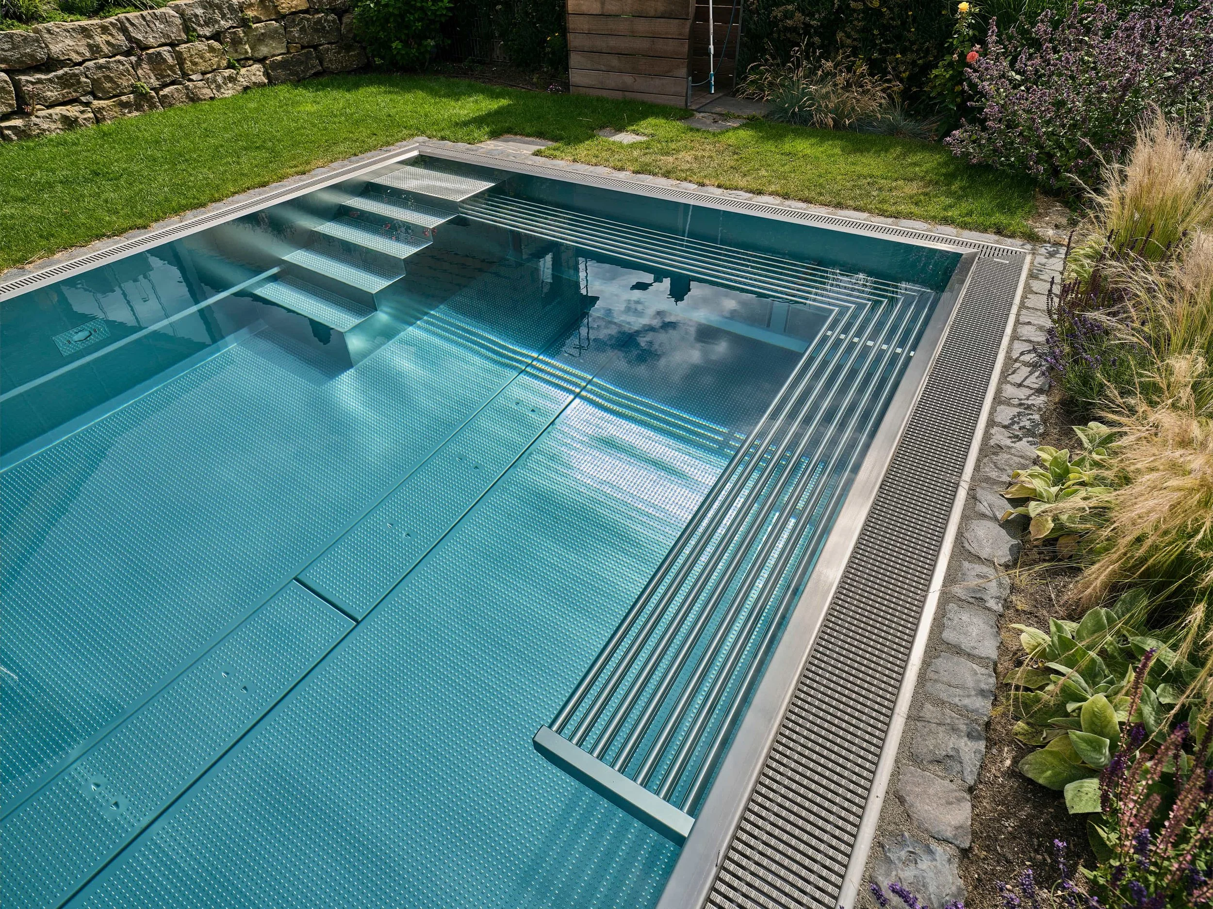 A modern rectangular swimming pool with metallic stairs, surrounded by grass, stone, and flowering plants in a backyard.