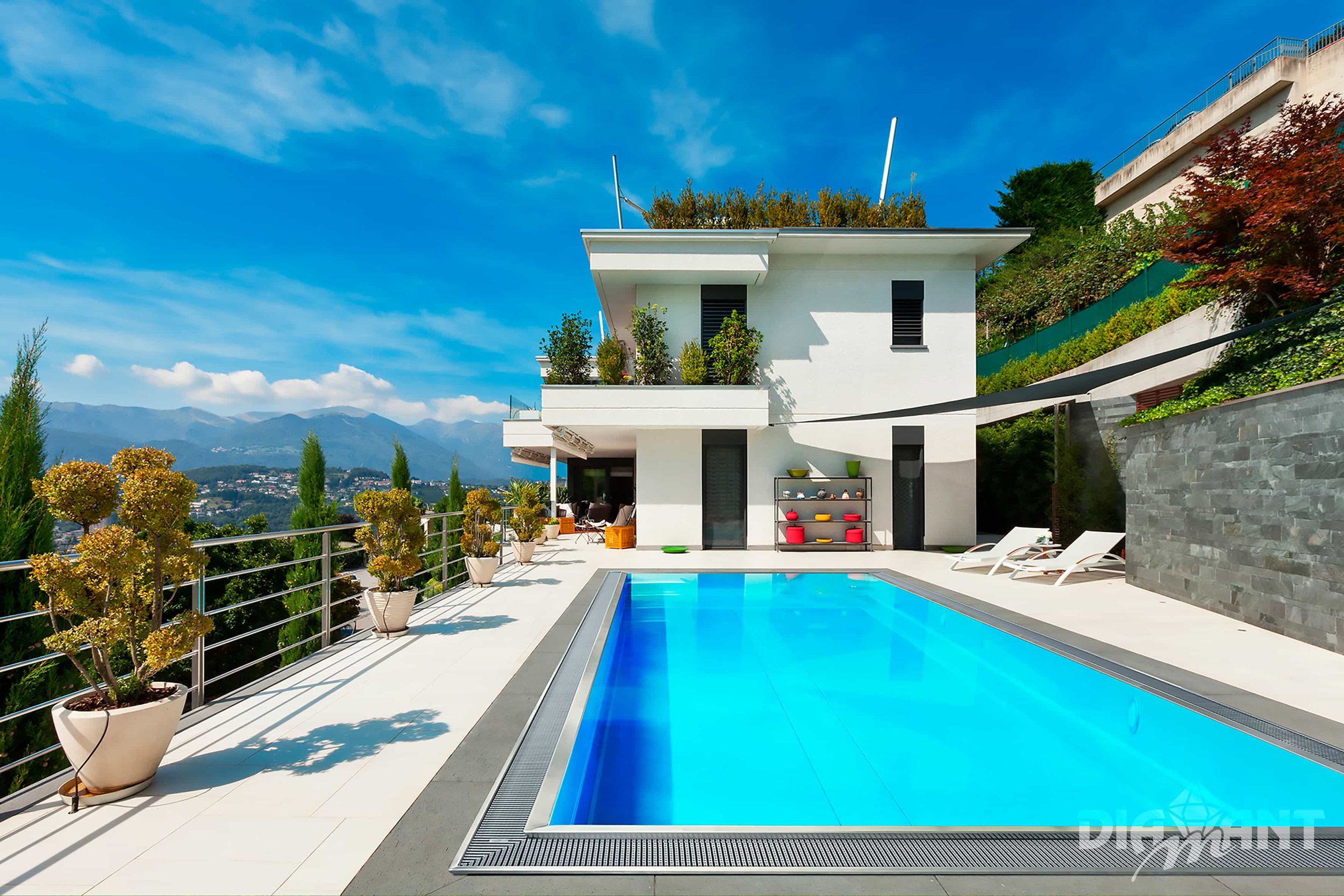 Modern house with a swimming pool on a hillside, surrounded by potted plants and lounge chairs, with mountains in the background and a partly cloudy blue sky.