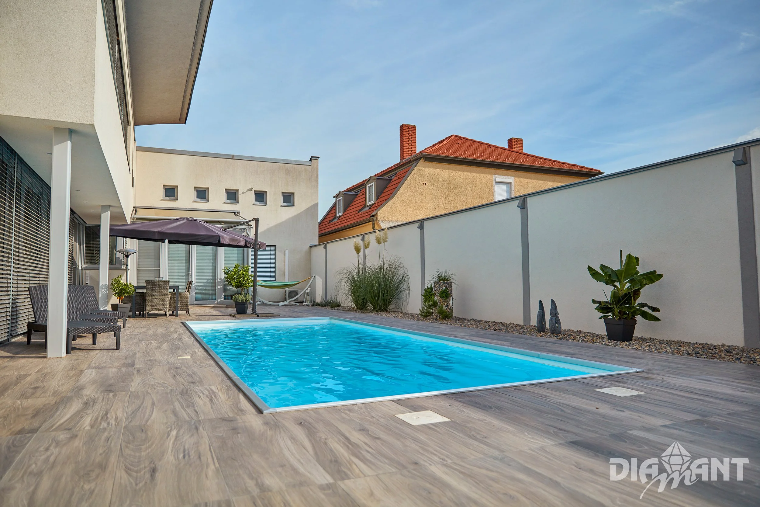 Residential backyard with a swimming pool, outdoor furniture, umbrella, potted plants, and a privacy fence under a clear blue sky.