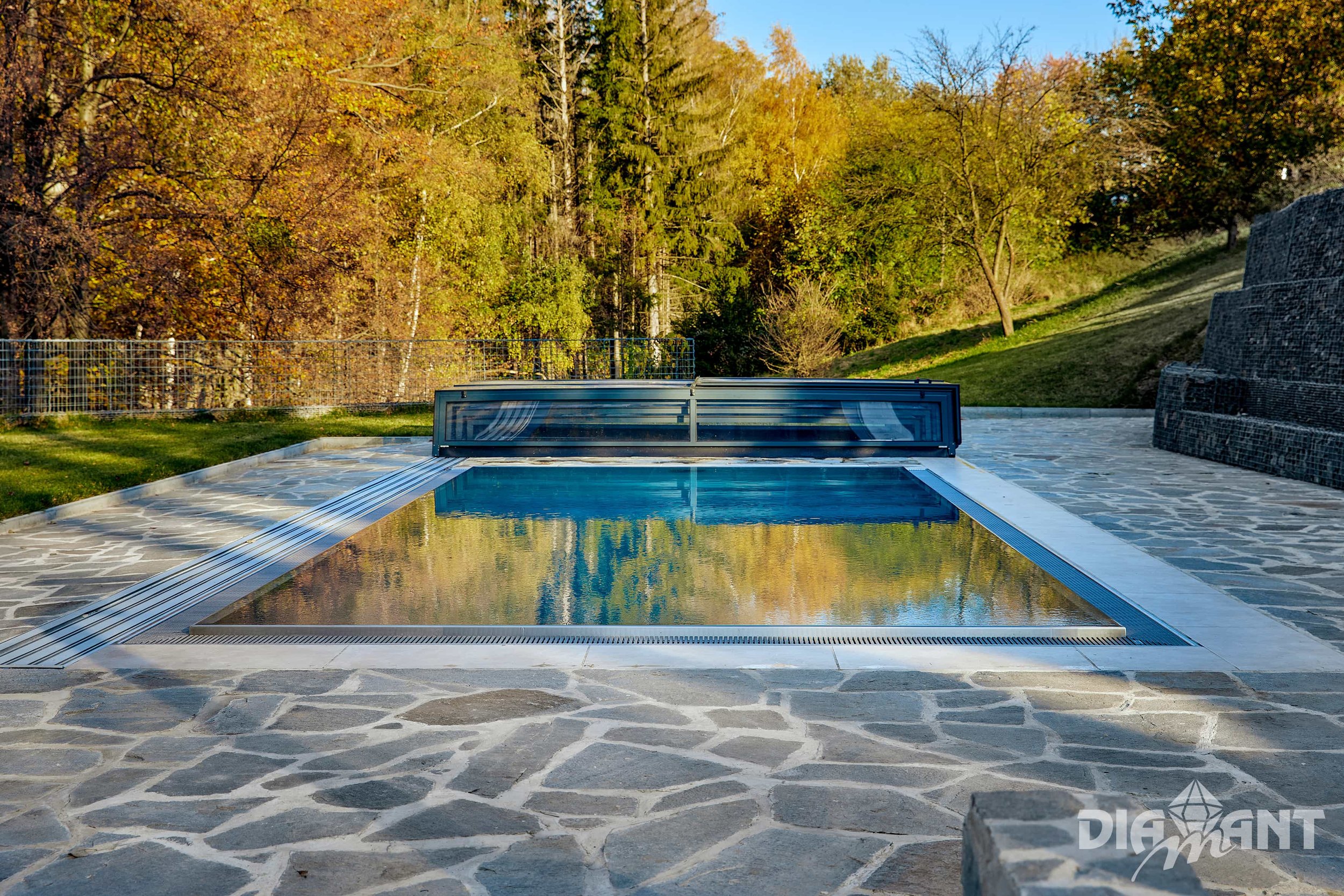 An outdoor swimming pool with a retractable cover, surrounded by a stone patio, with trees and a hillside in the background, on a sunny day.