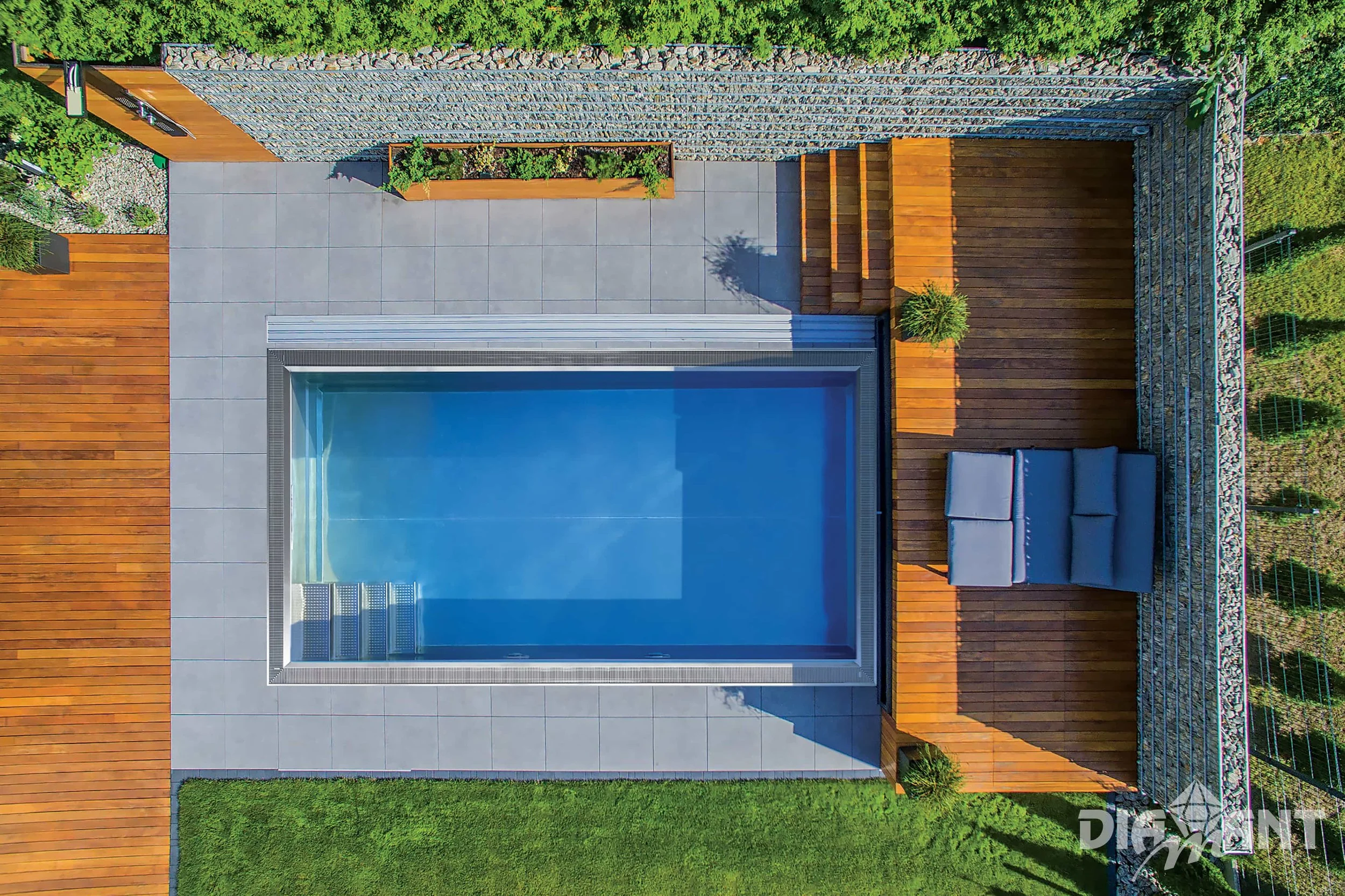 A modern backyard with a rectangular swimming pool, surrounded by a tiled area and wooden decking. There are stairs leading into the pool, and outdoor furniture, including a sofa and some potted plants, on the deck. The yard is enclosed with a stone wall and greenery.