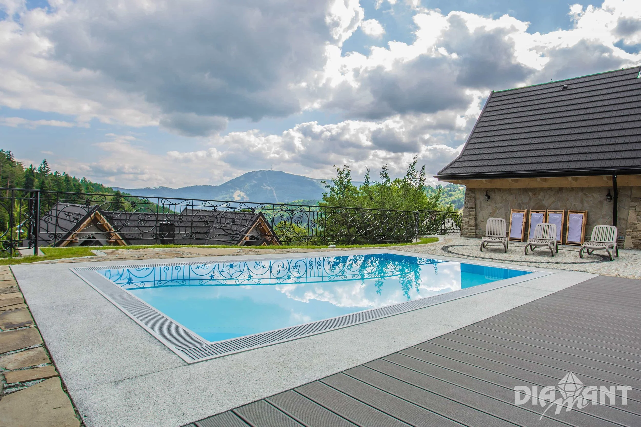 Outdoor swimming pool reflecting cloudy sky, surrounded by wood and stone deck chairs, with mountains and trees in the background.