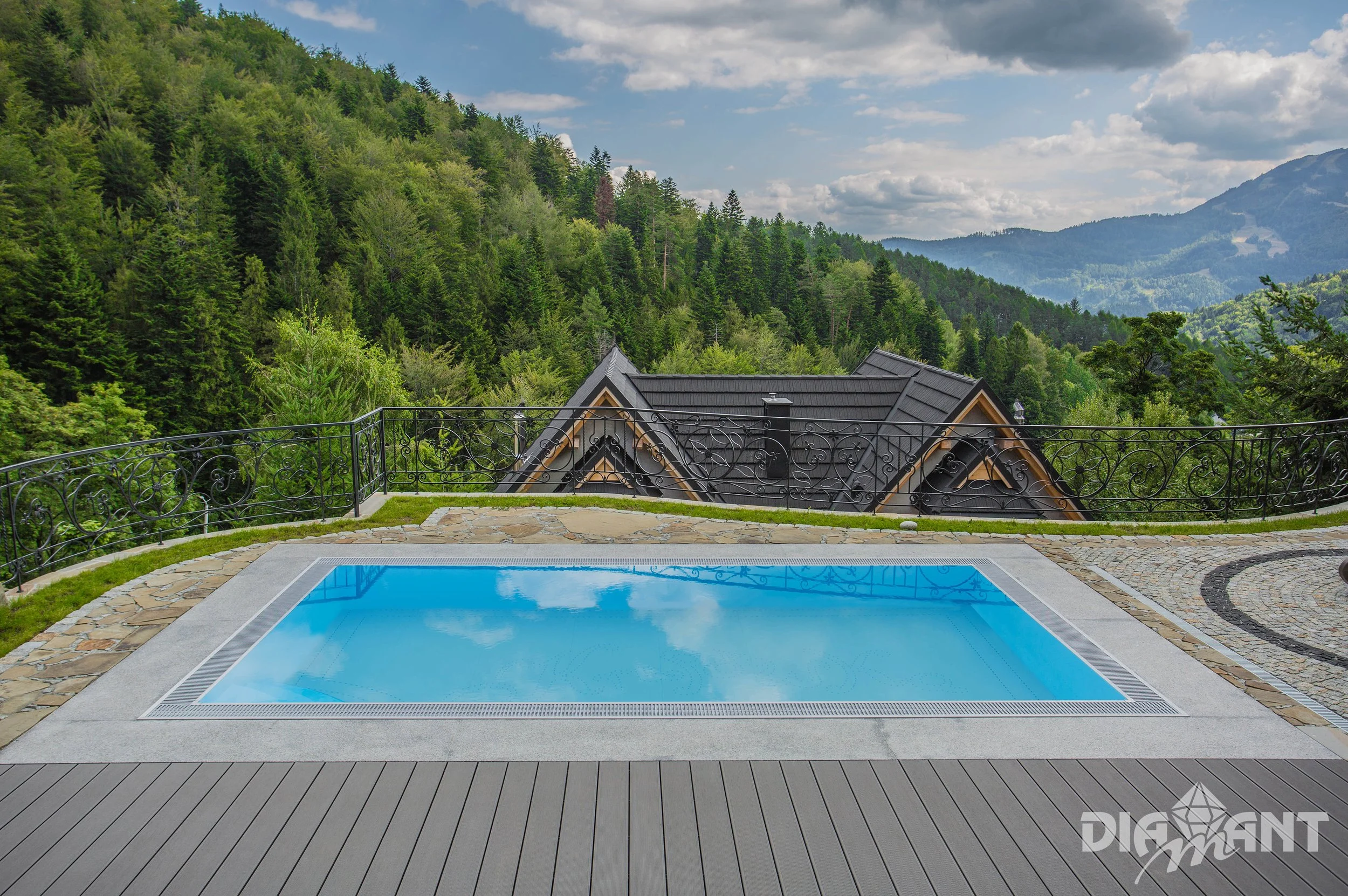 Small swimming pool on a terrace with a mountain view, in a natural setting with lush green trees, cloudy sky, and rooftops of houses in the background.