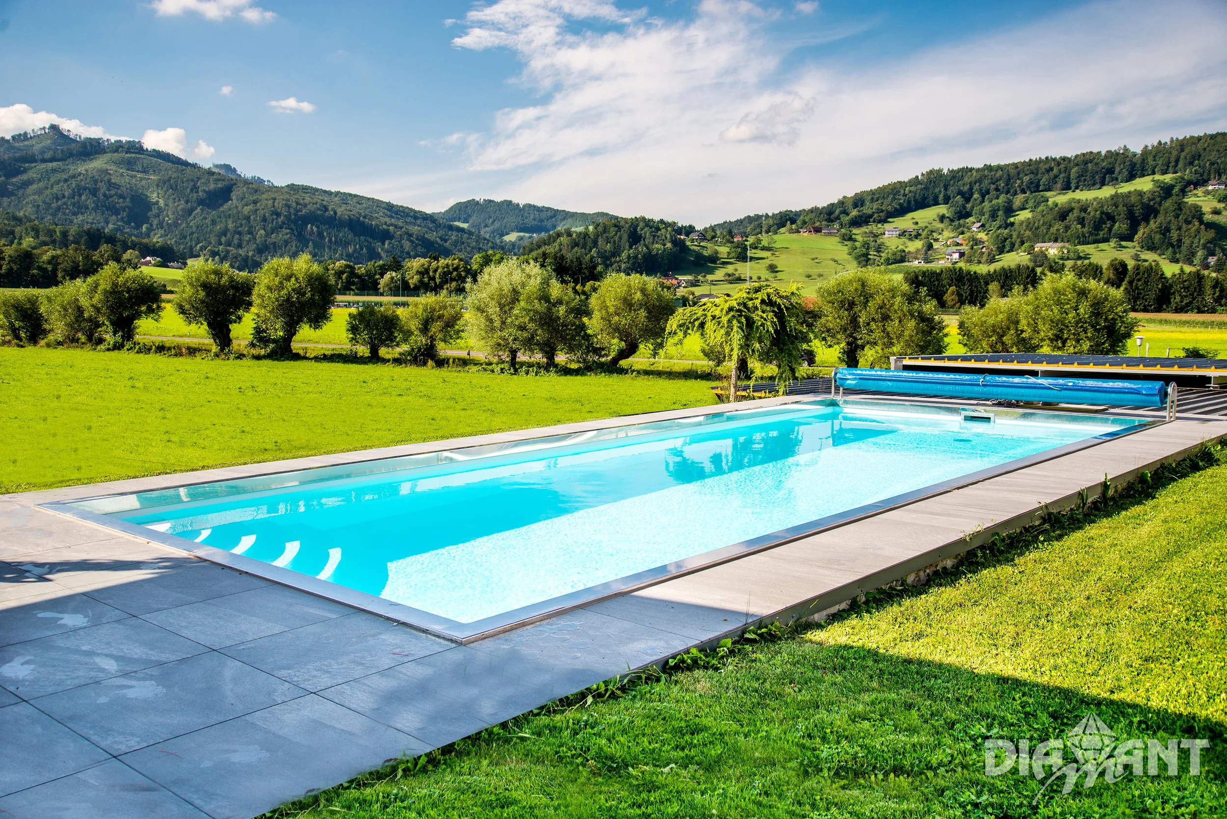 A rectangular outdoor swimming pool with clear blue water situated on a lush green lawn, surrounded by trees and rolling hills under a partly cloudy sky.