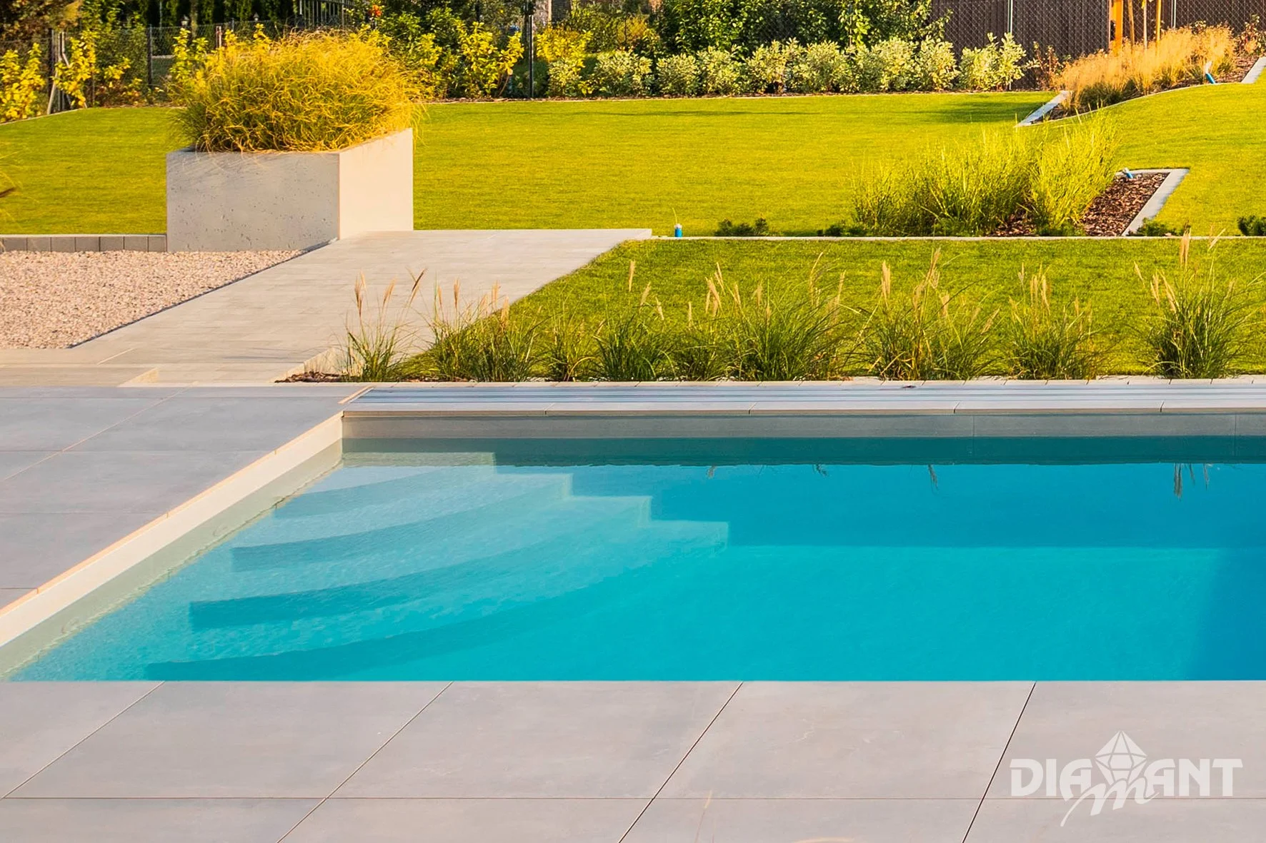 A backyard swimming pool with a surrounding tiled deck, green grass, and landscaped plants, with a background of trees and shrubbery.